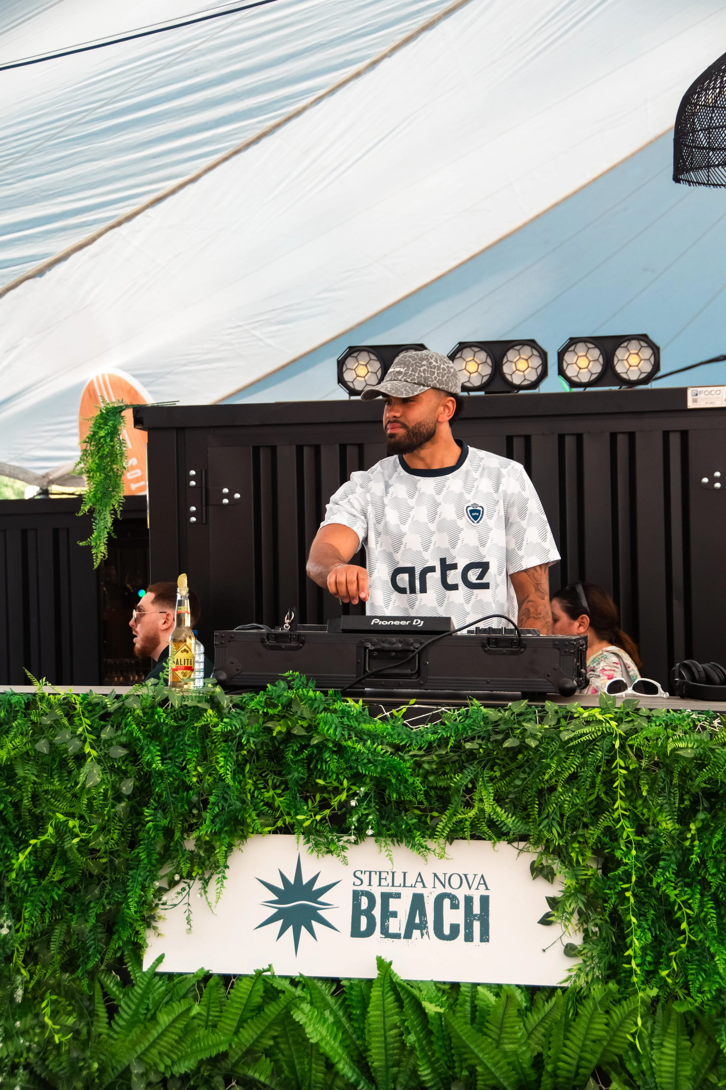 A male DJ wearing a patterned cap and white shirt with logos, standing behind DJ equipment, at Stella Nova Beach event, surrounded by greenery, with a large sign reading Stella Nova Beach in front.
