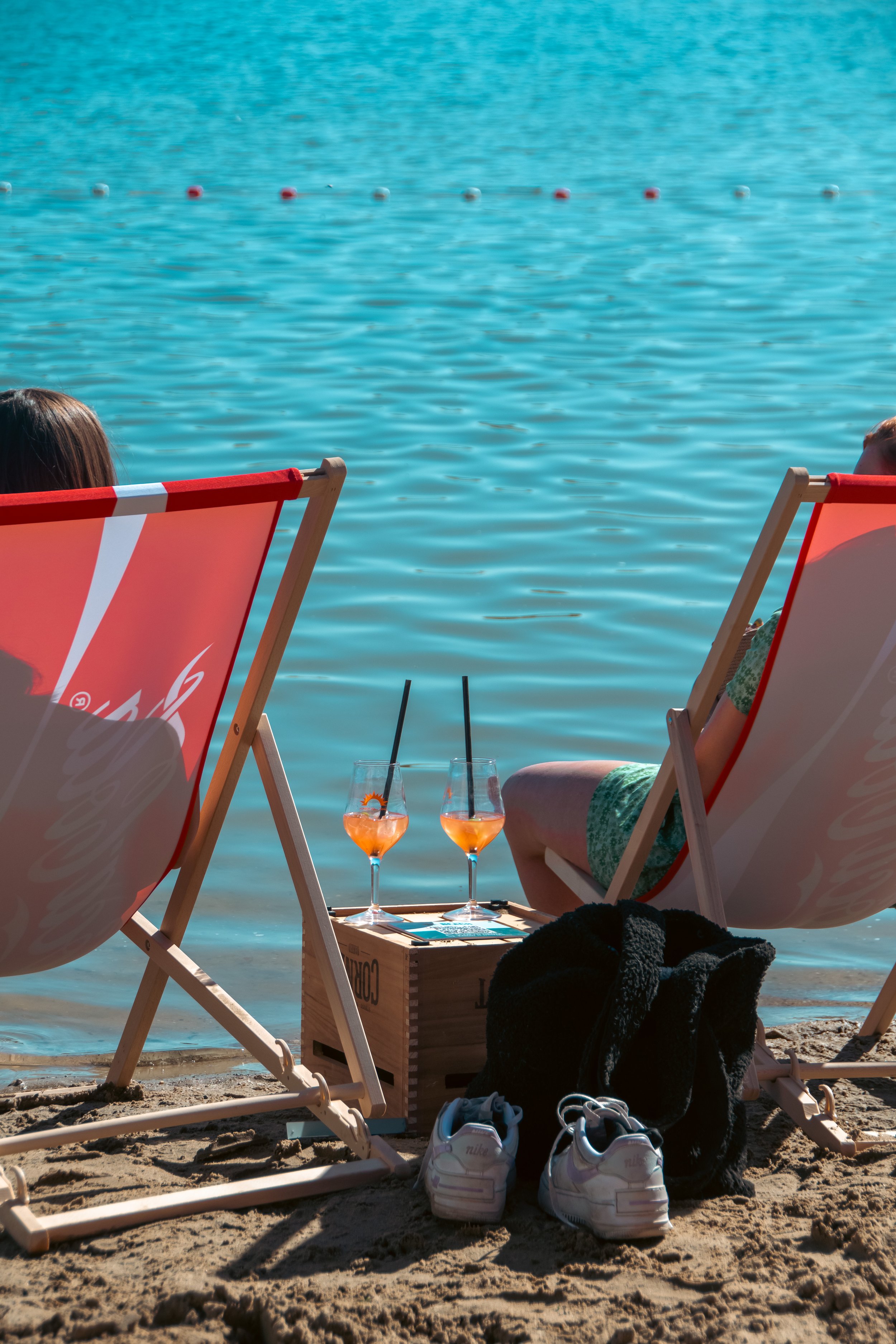 Two women relax on beach chairs by the water, facing the ocean. Two glasses with orange drinks and black straws sit on a wooden crate between them. Various shoes and a black towel are on the sandy ground.