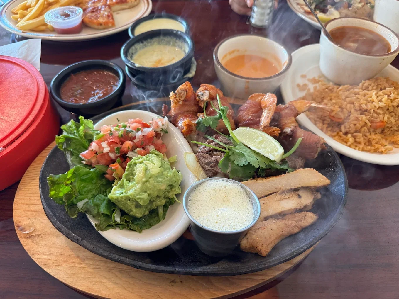 A traditional Mexican meal served on a hot stone platter, featuring guacamole, salsa, grilled meats, and lime, with Mexican rice and various sauces on side dishes.