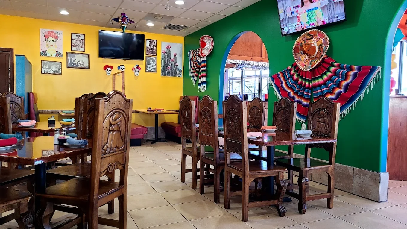 Interior of a colorful Mexican restaurant with yellow and green walls, decorated with Frida Kahlo artwork, papel picado, a sombrero, and a vibrant textile wall hanging. Wooden tables and chairs are arranged with place settings.