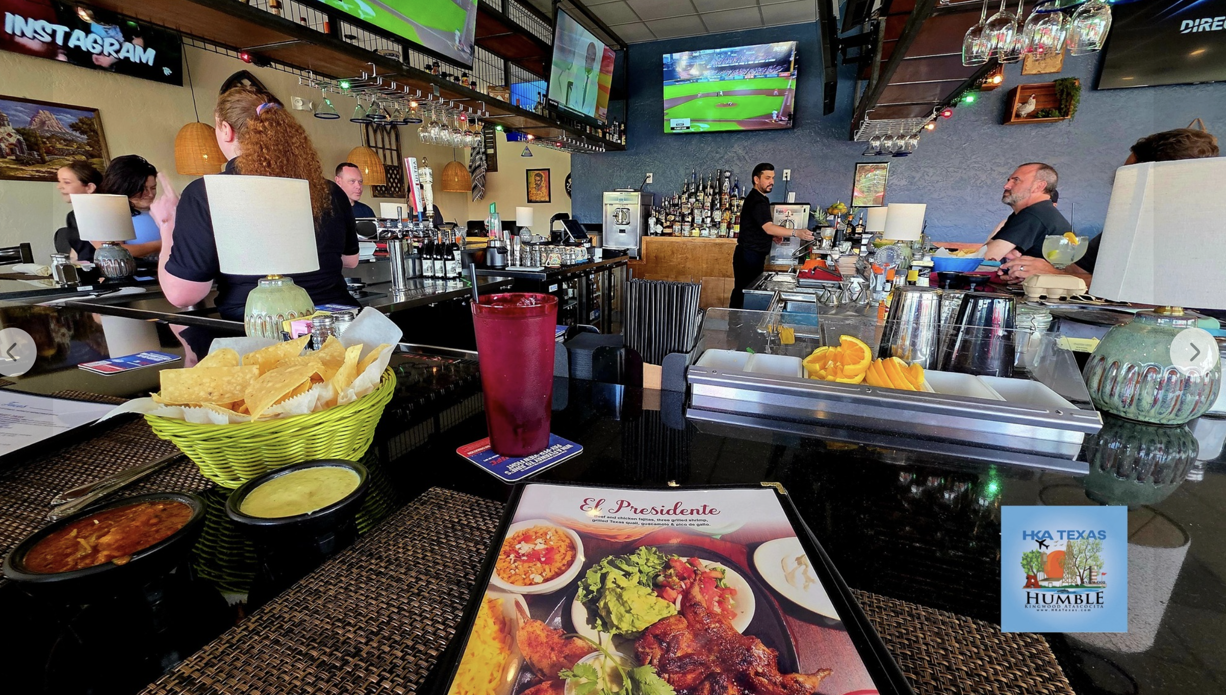 Interior of a restaurant with a bar area, people seated at tables, several large TV screens showing a baseball game, and a bartender preparing drinks. On the table in the foreground, there is a menu, a red drink, chips with two dips, and a tray with sliced fruit.