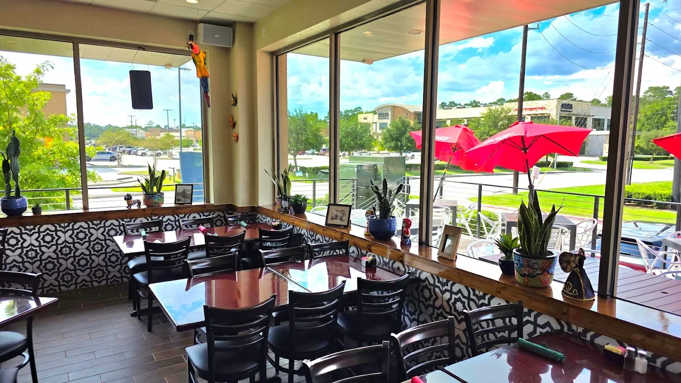 Interior of a restaurant with black chairs and tables, decorative plant pots, and large windows showing an outdoor patio with red umbrellas and a sunny day.