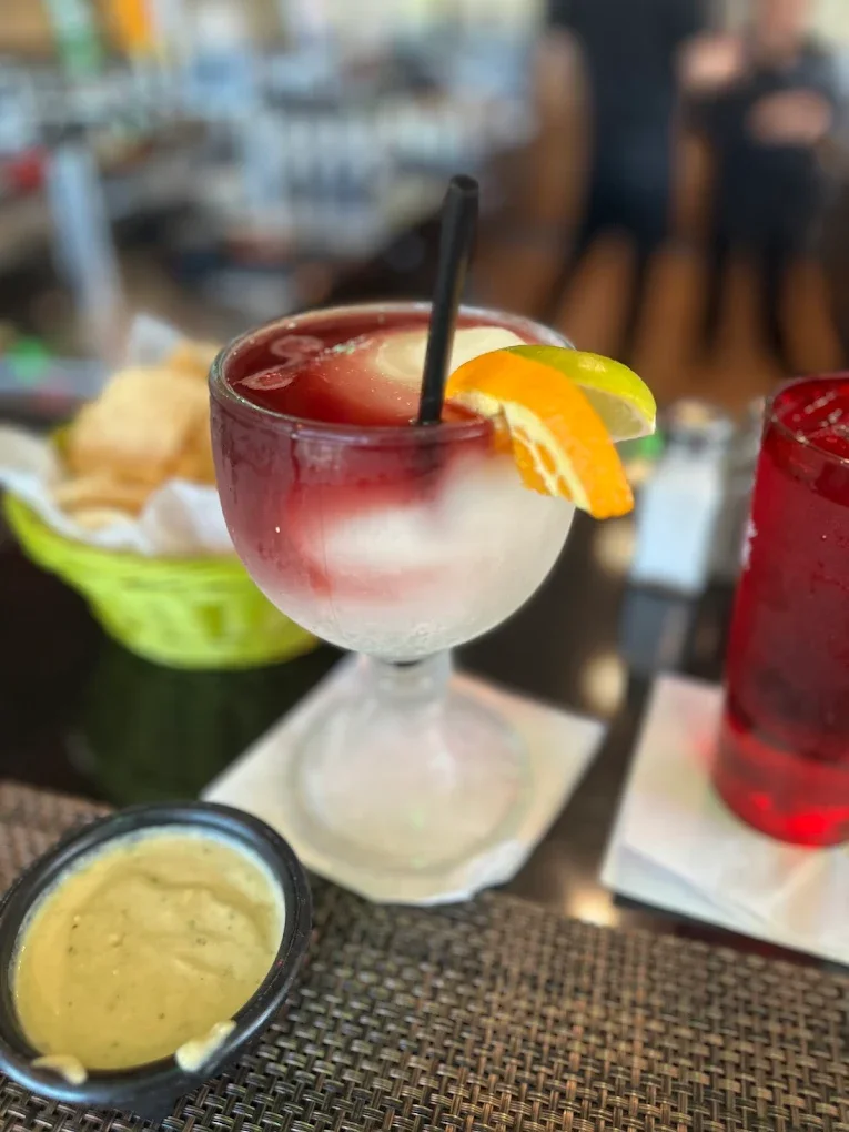 A cocktail with red and white layers garnished with a lemon and lime wedge, served in a stemmed glass with a black straw, on a woven placemat at a restaurant table.