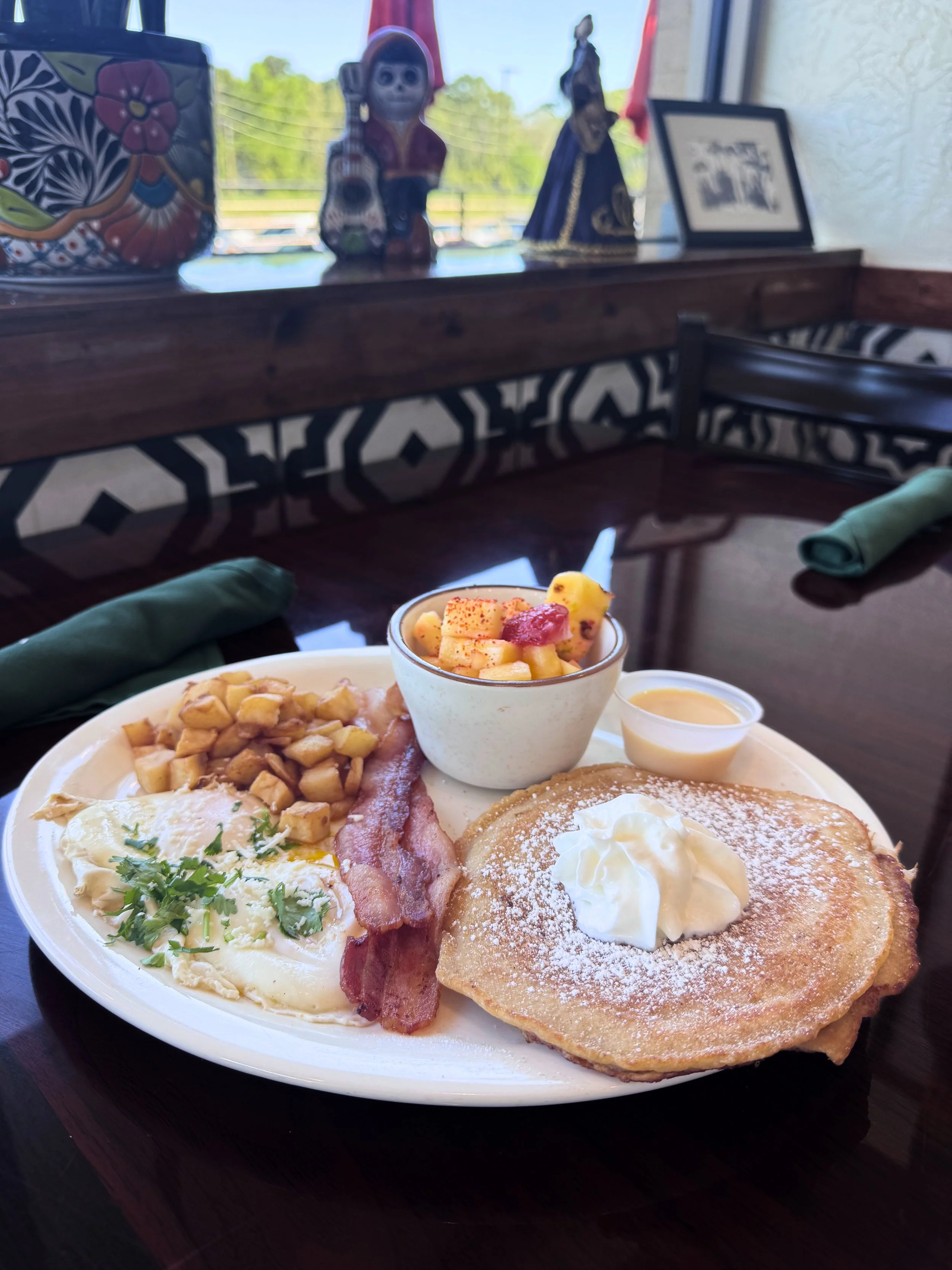 A plate of breakfast food including pancakes with whipped cream, scrambled eggs with herbs, bacon, breakfast potatoes, and fruit salad with apple slices and strawberries, served with syrup and butter, on a dark wooden table.