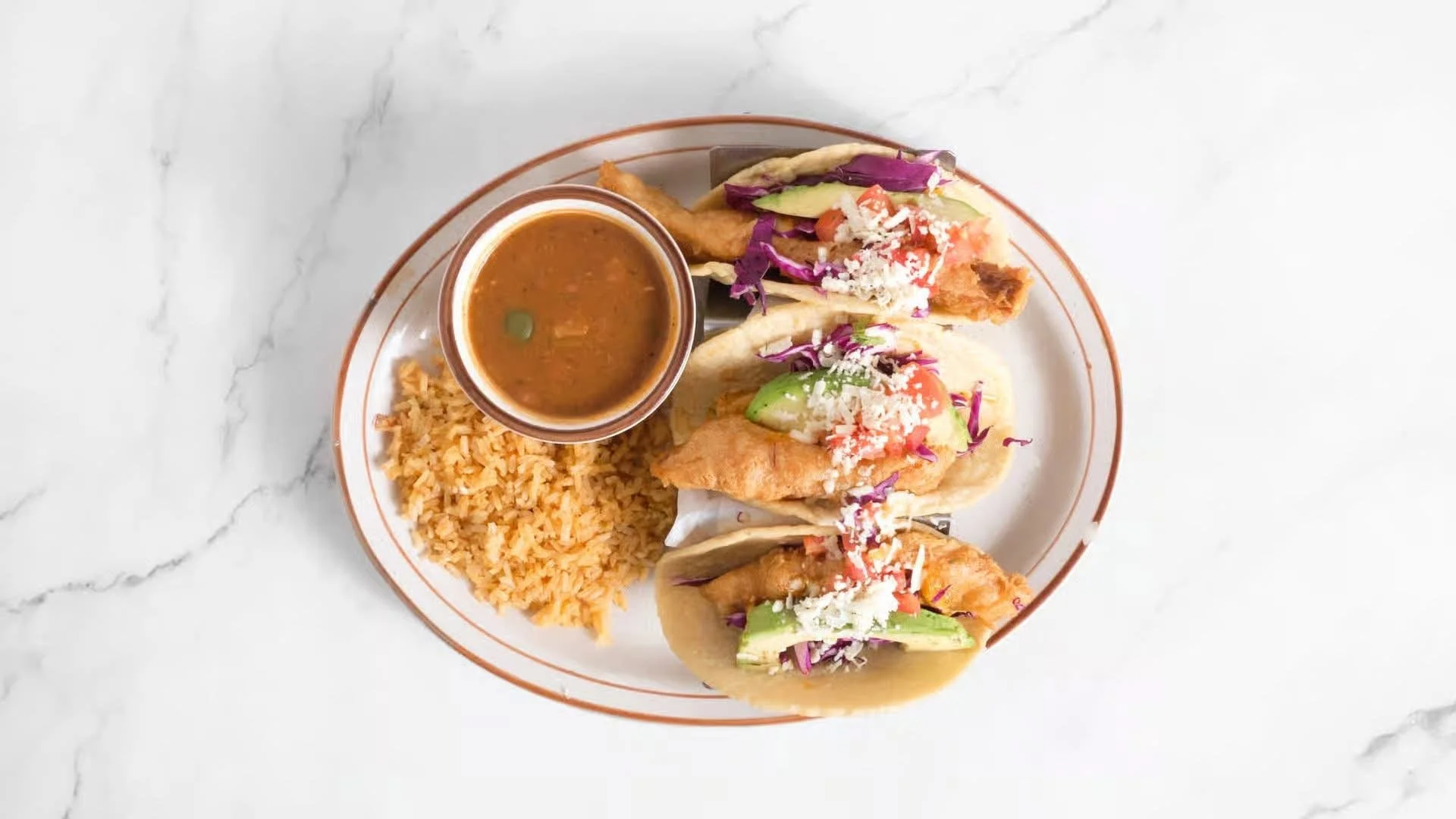 Plate of three fish tacos topped with shredded cheese, cabbage, and vegetables, served with Mexican rice and a small bowl of sauce on a white marble surface.