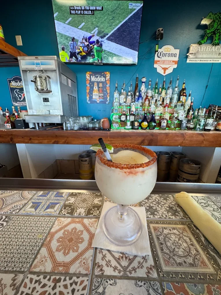 A large, frozen margarita with salted rim, lime wedge, and straw on a decorative tiled bar counter in a bar setting with bottles and TV in background