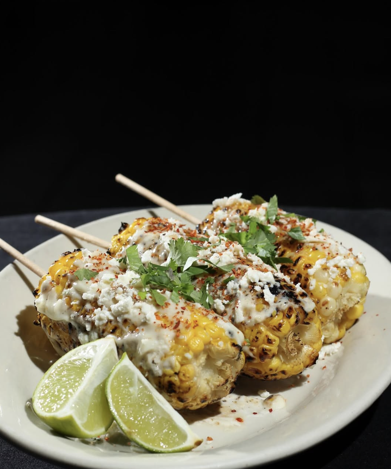 Three grilled corn on the cob skewers topped with mayonnaise, cheese, chili powder, and cilantro, garnished with lime wedges on a white plate.