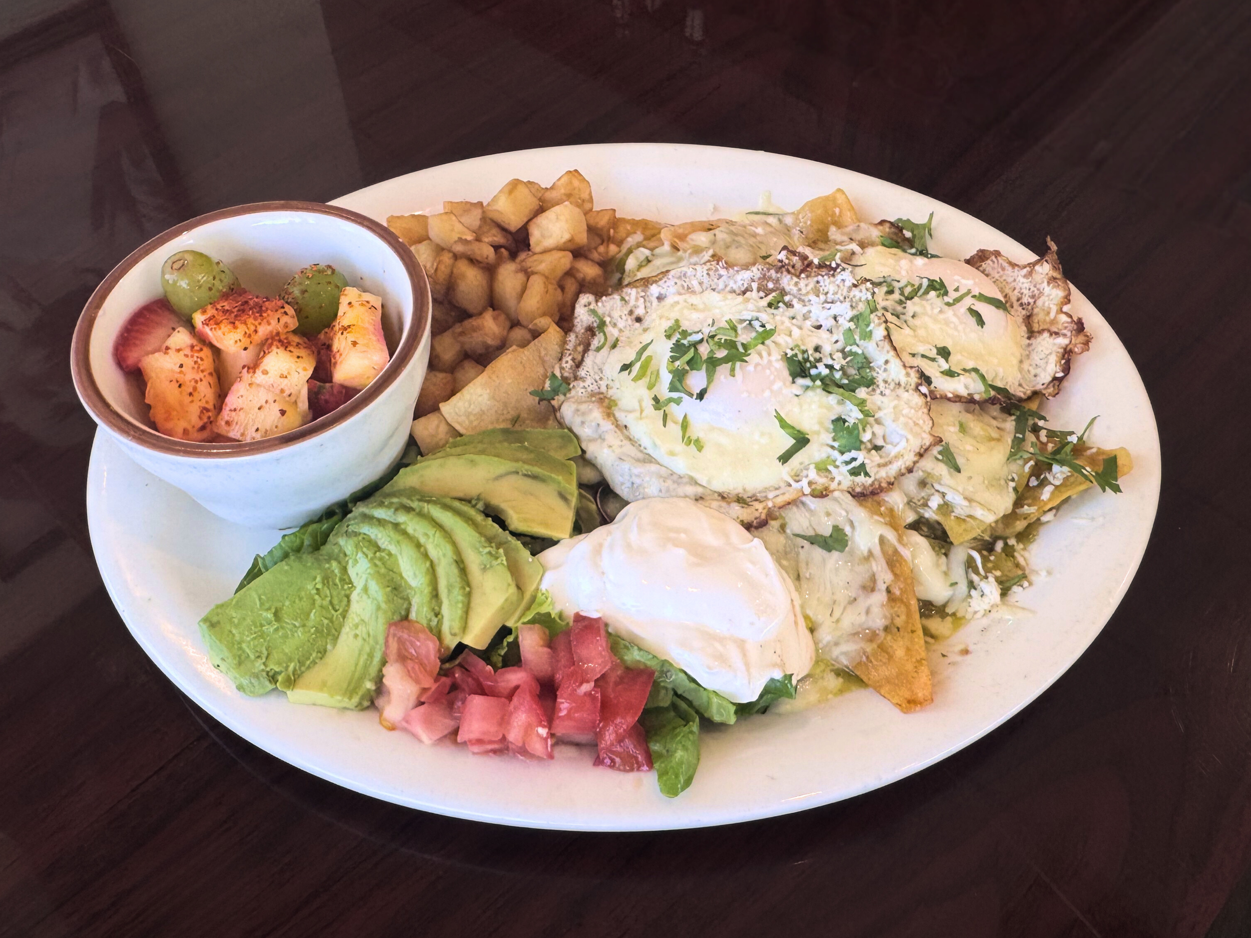 Plate with fried eggs, avocado slices, diced tomatoes, fried potatoes, hash browns, and a side of fruit salad with grapes, strawberries, and pineapple