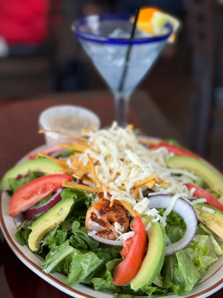 Fresh green lettuce, sliced tomatoes, avocado, onion, and cheese on a plate with fried shrimp and guacamole, with a cocktail in a martini glass in the background.