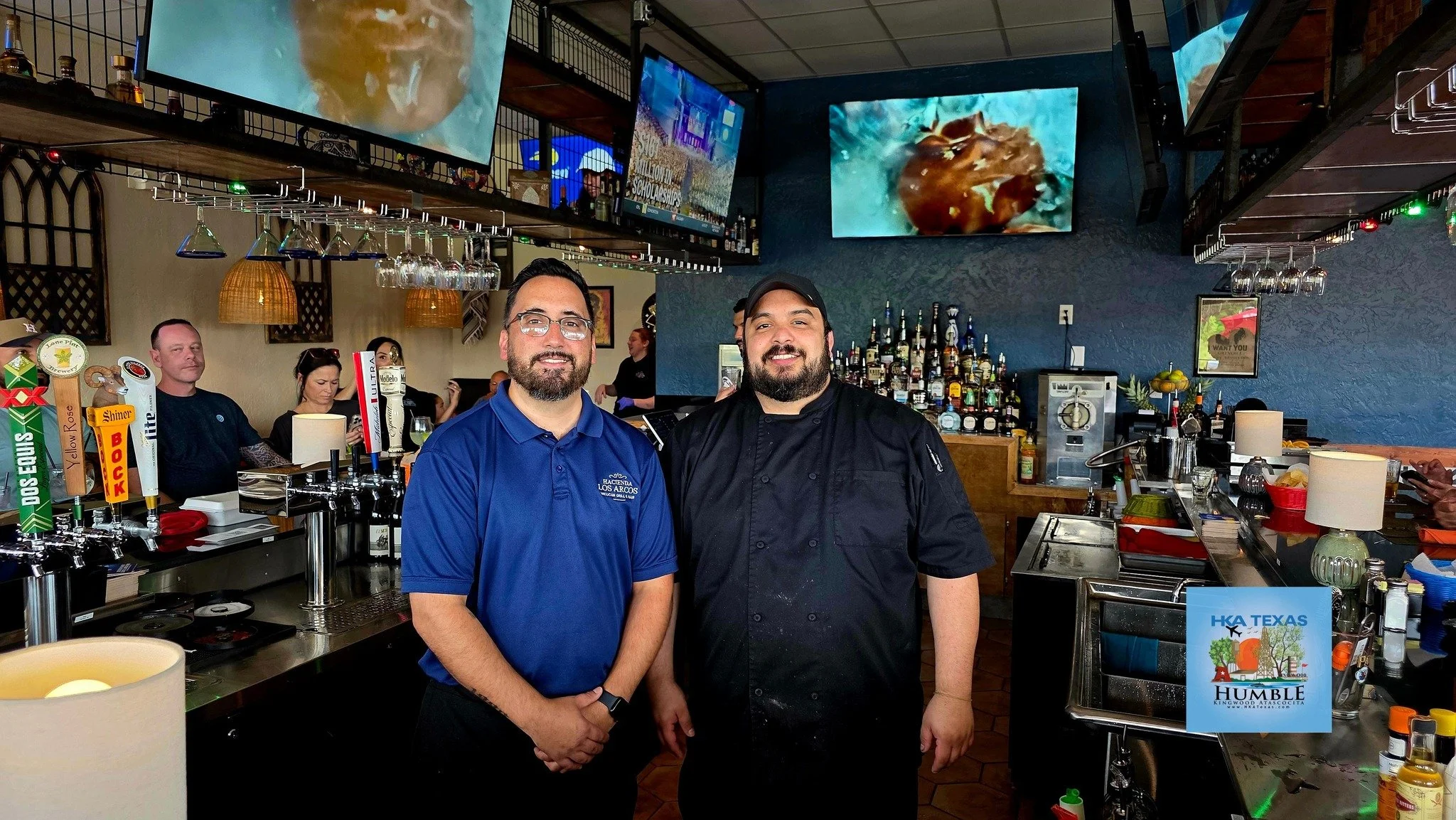 Two men standing behind a bar counter in a restaurant or bar. One is wearing a blue polo shirt with a logo, and the other is dressed in a black chef's coat and cap. There are multiple televisions mounted on the wall behind them, and various bottles of alcohol are arranged on shelves. Other patrons are visible in the background.