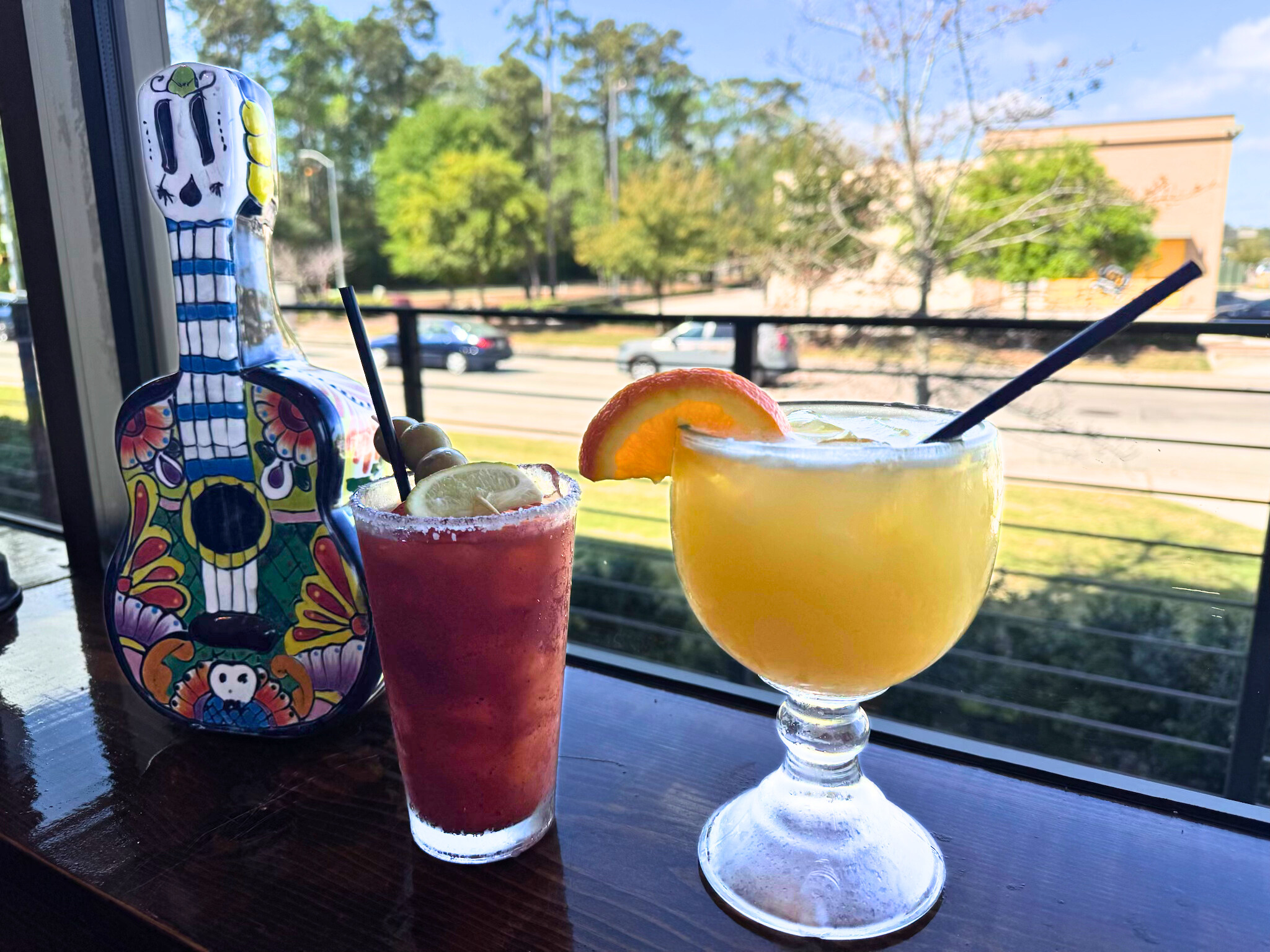 Two colorful cocktails on a dark wooden table next to a decorative skeleton figurine, with a background of green trees, and a building.