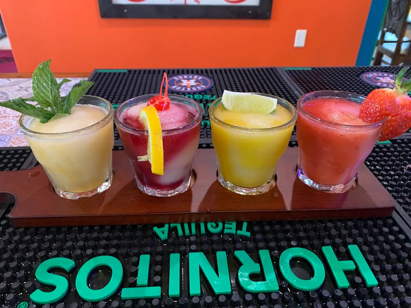 Four colorful cocktails on a wooden serving board at a bar, topped with a mint sprig, cherry and lemon, lemon wedge, strawberry, and lime wedge respectively.