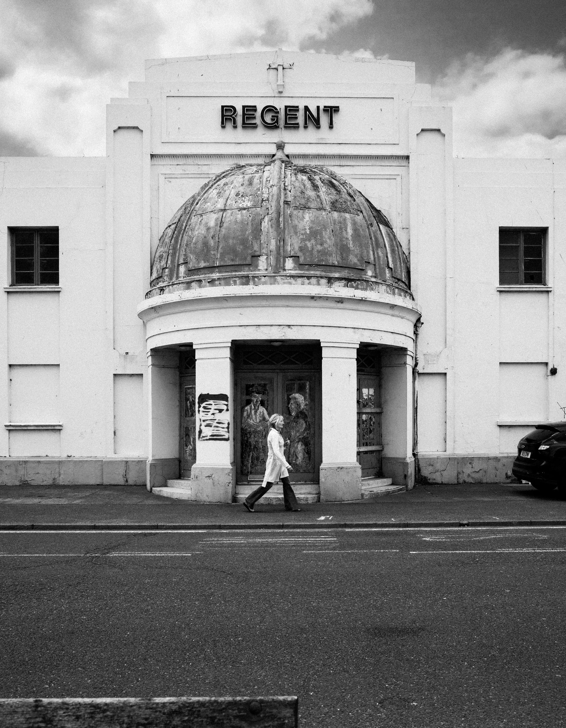 Old theater building with a weathered dome and the sign 'Regent' on the front, featuring a woman walking past on the sidewalk.