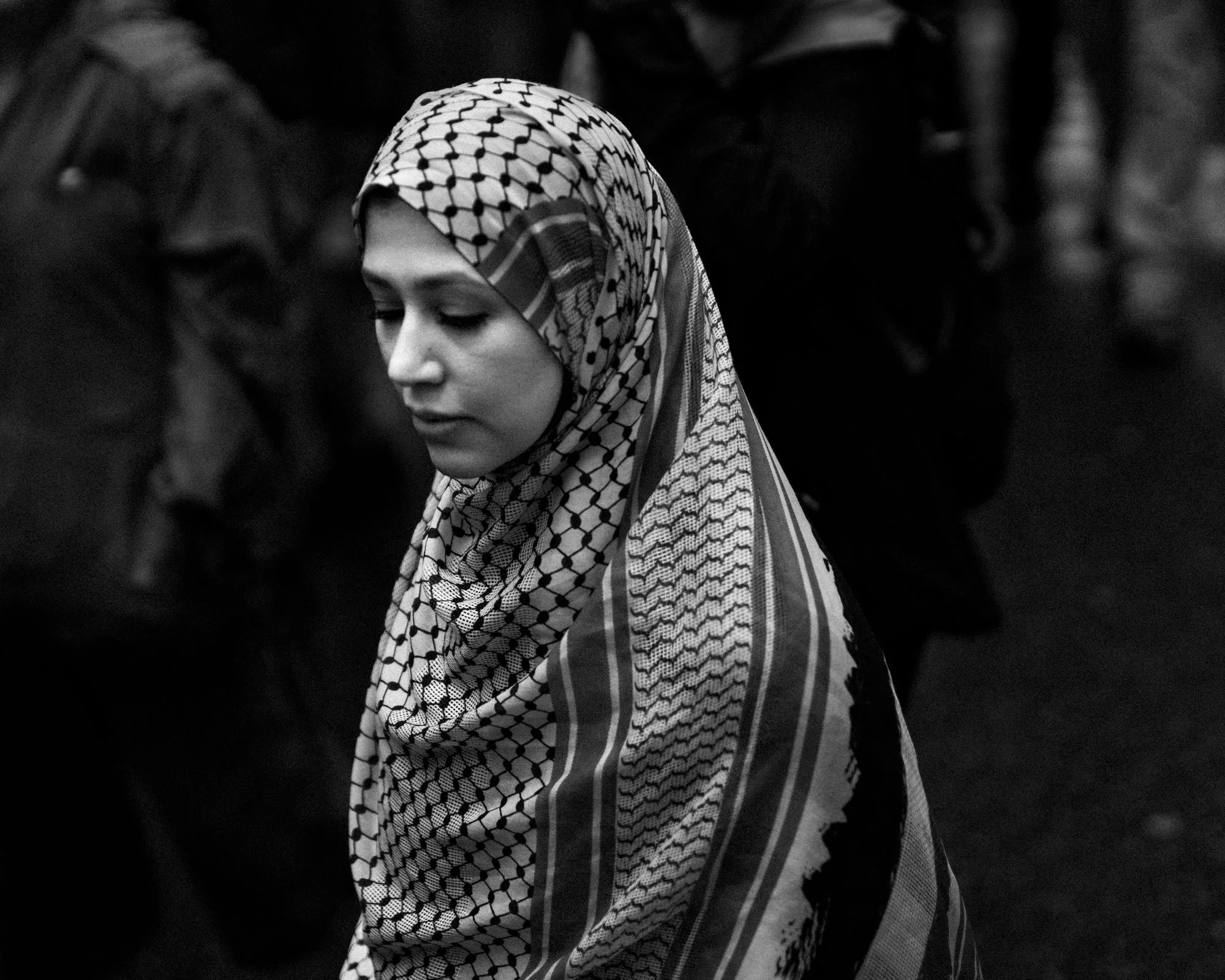 A young woman with a patterned headscarf looking downward in a crowd.