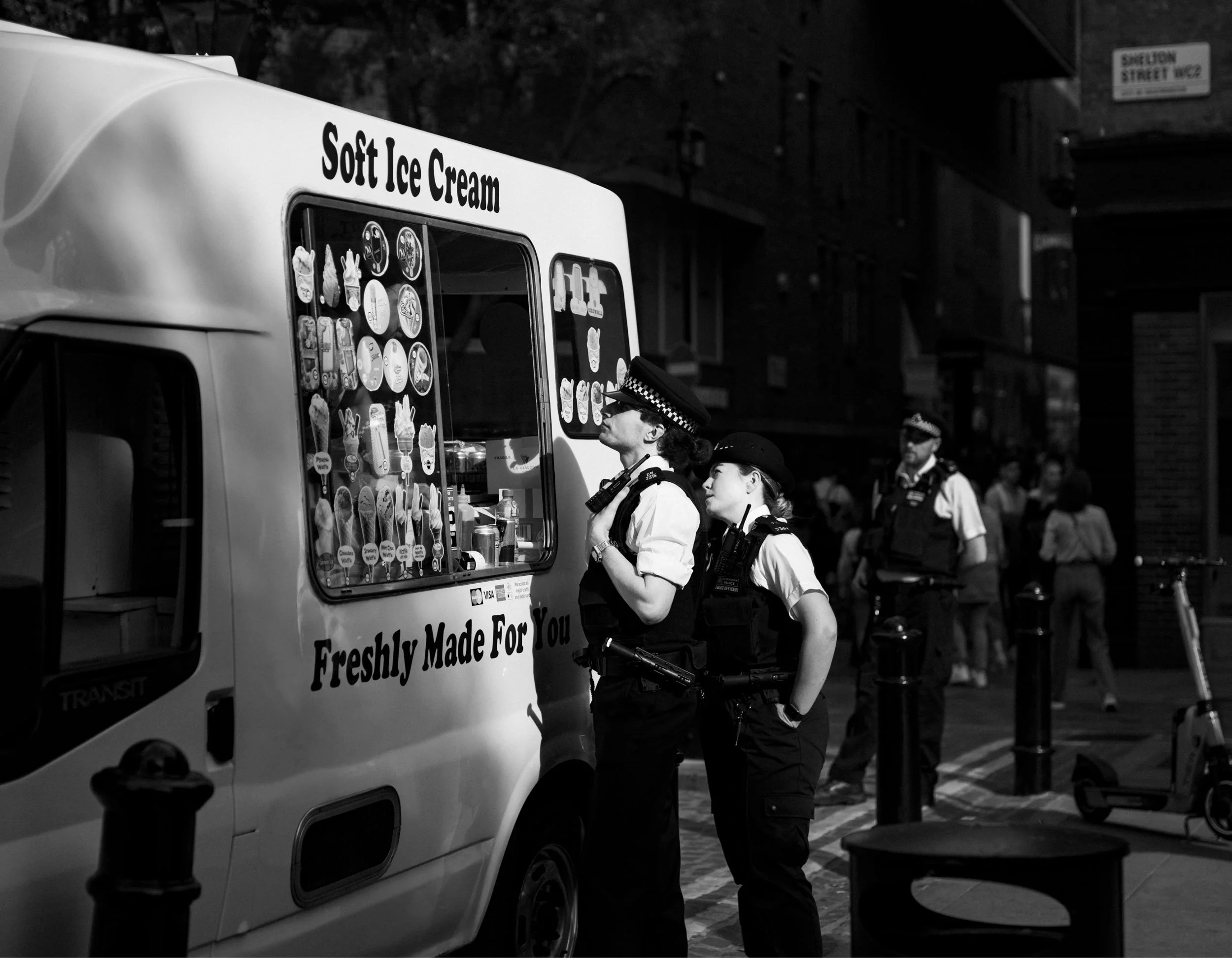 Two police officers stand in front of an ice cream truck on a city street at night. The truck sells soft ice cream and has various ice cream images on the window.