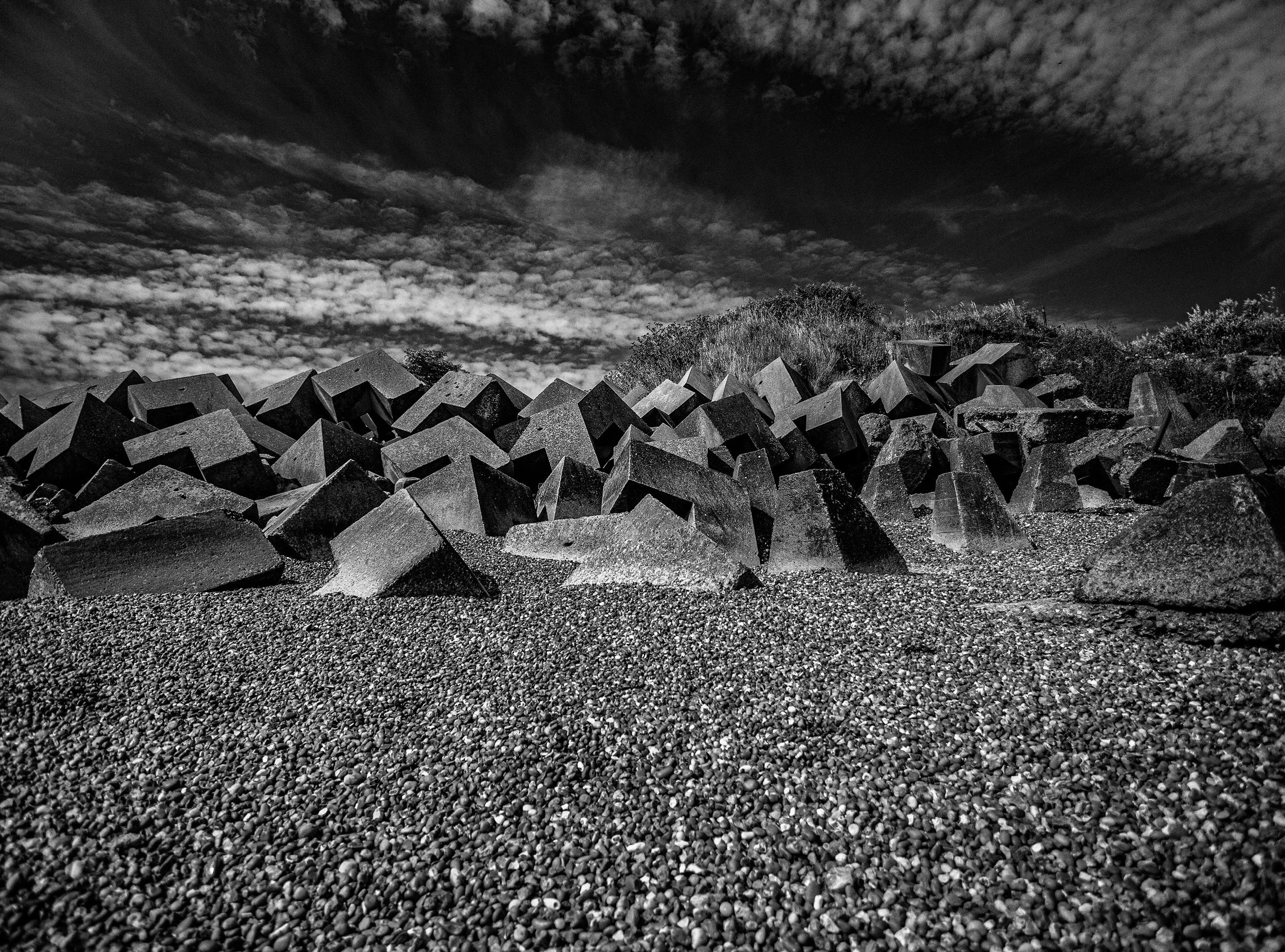 Black-and-white photograph of a large pile of concrete blocks and rocks under a cloudy sky.