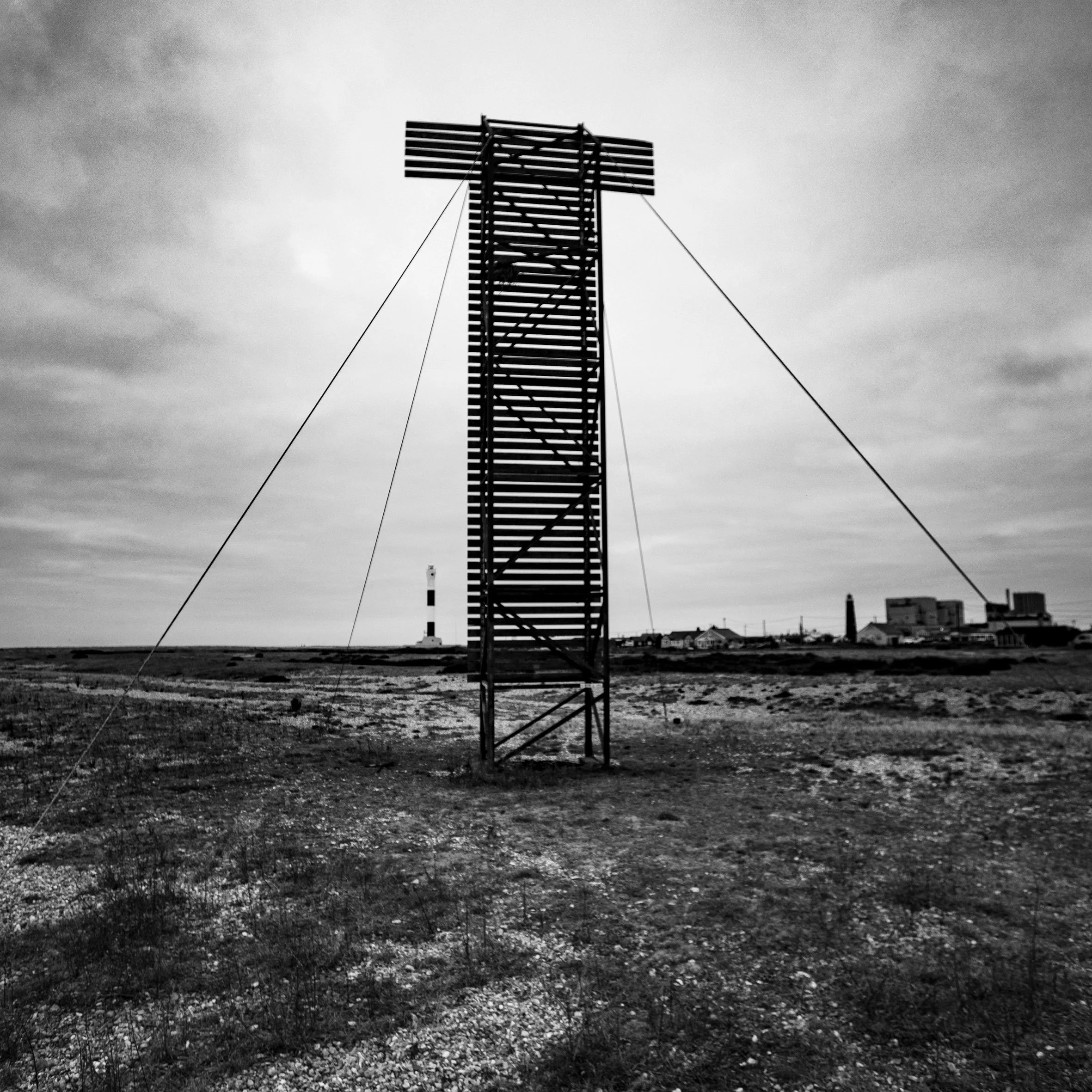 Black and white photo of a tall wooden structure supported by guy wires on an open barren landscape, with a lighthouse and buildings in the background under a cloudy sky.