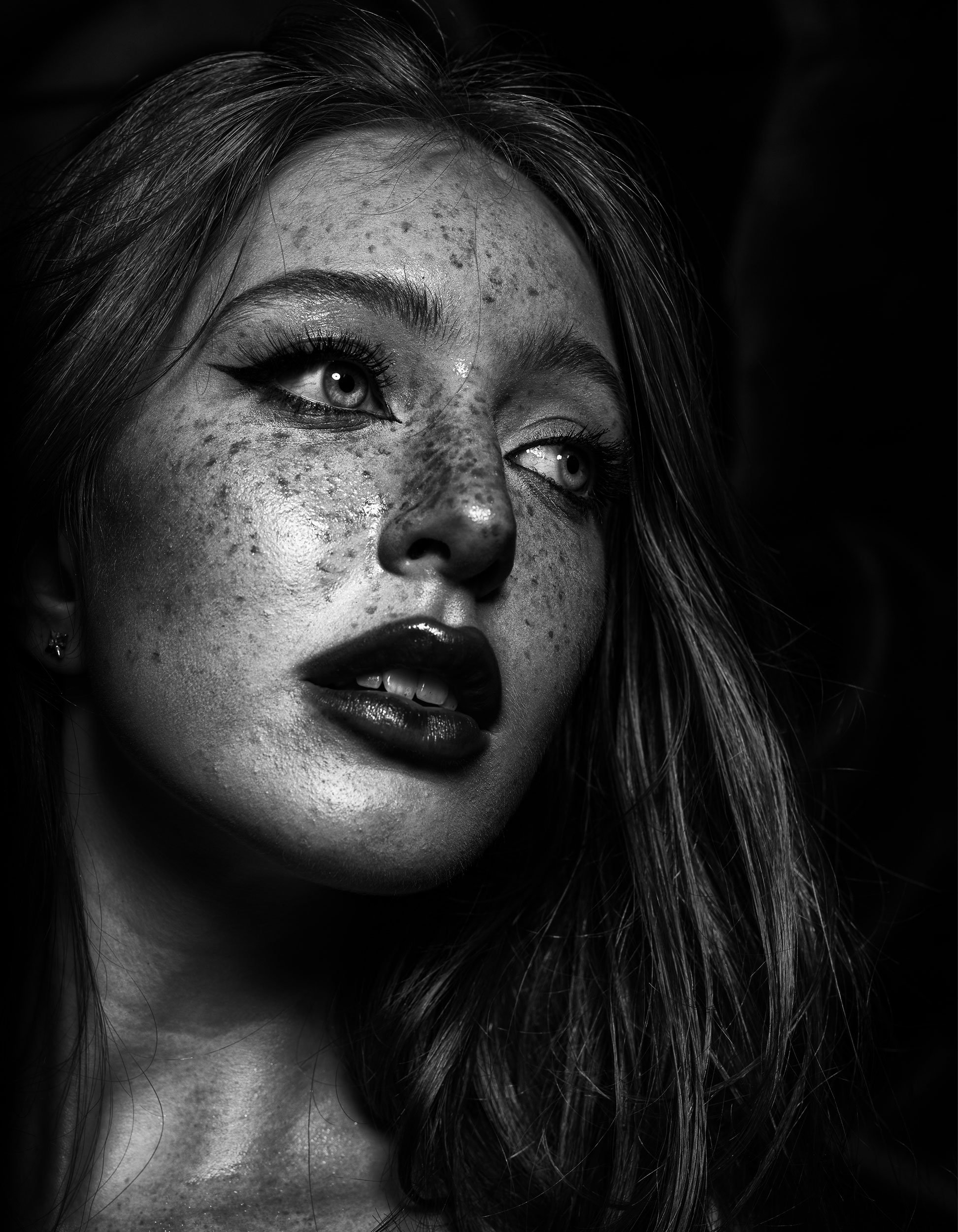 Black and white close-up portrait of a young woman with freckles, dramatic eye makeup, and dark lipstick, looking slightly upward.