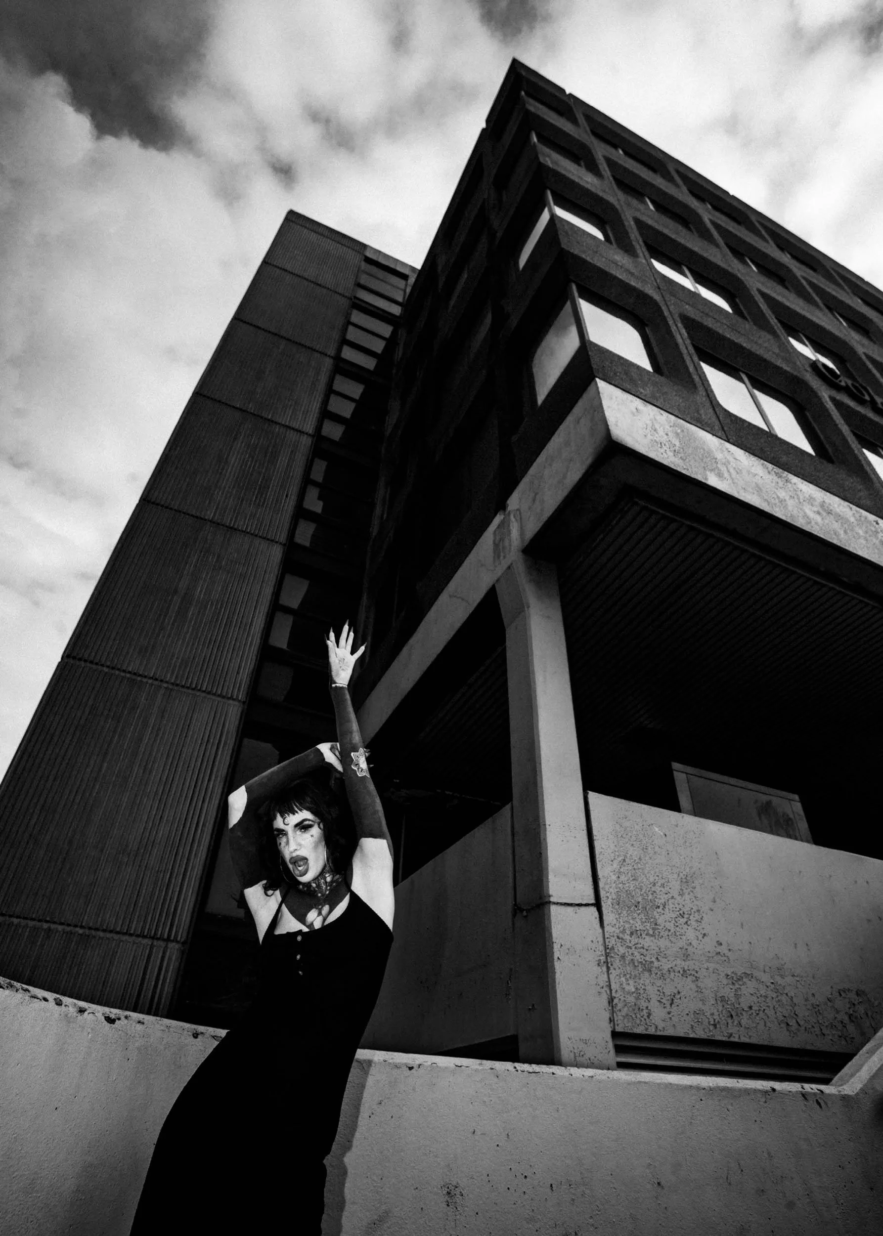 Black and white photo of a woman with dark hair and makeup, wearing a black dress with gloves, posing with one arm raised in front of a tall modern building under a cloudy sky. Chloe Rank-Harrington