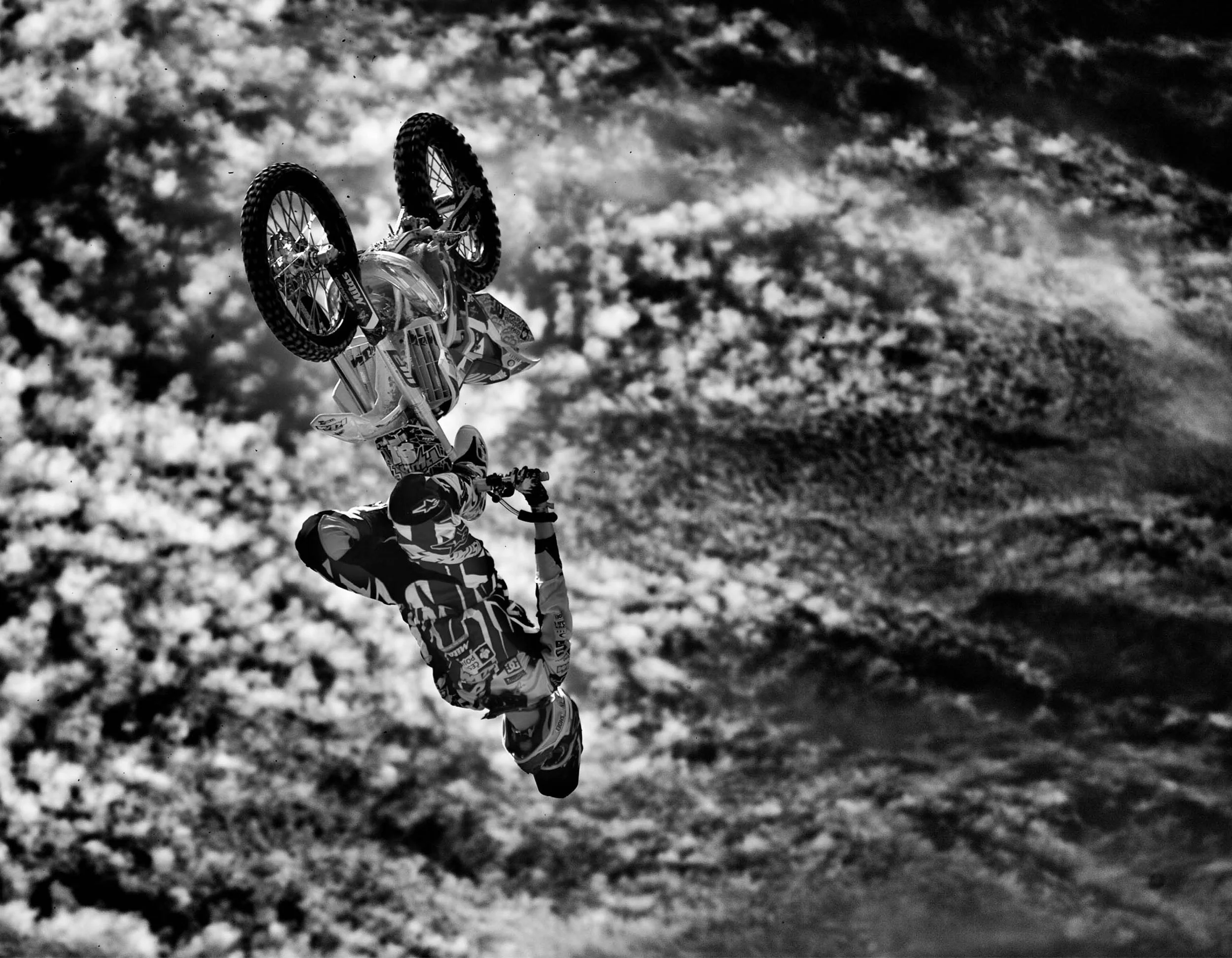 Black and white photo of a motocross rider performing a flip in mid-air against a cloudy sky background.