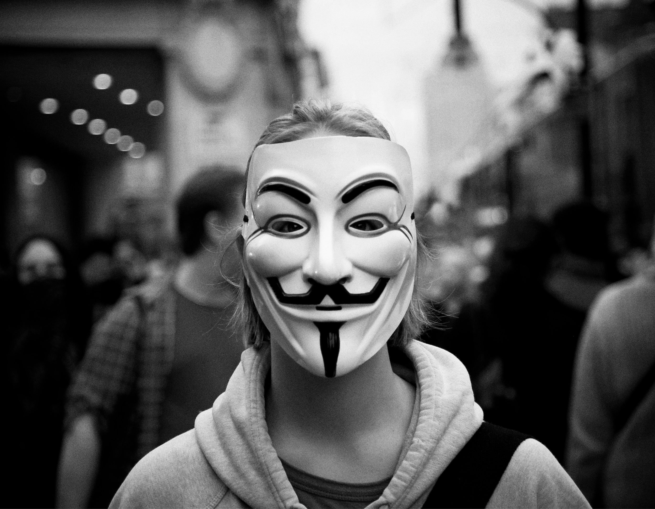 A person wearing a Guy Fawkes mask, hoodie, and gloves stands in a line of similar masked individuals in an urban setting.