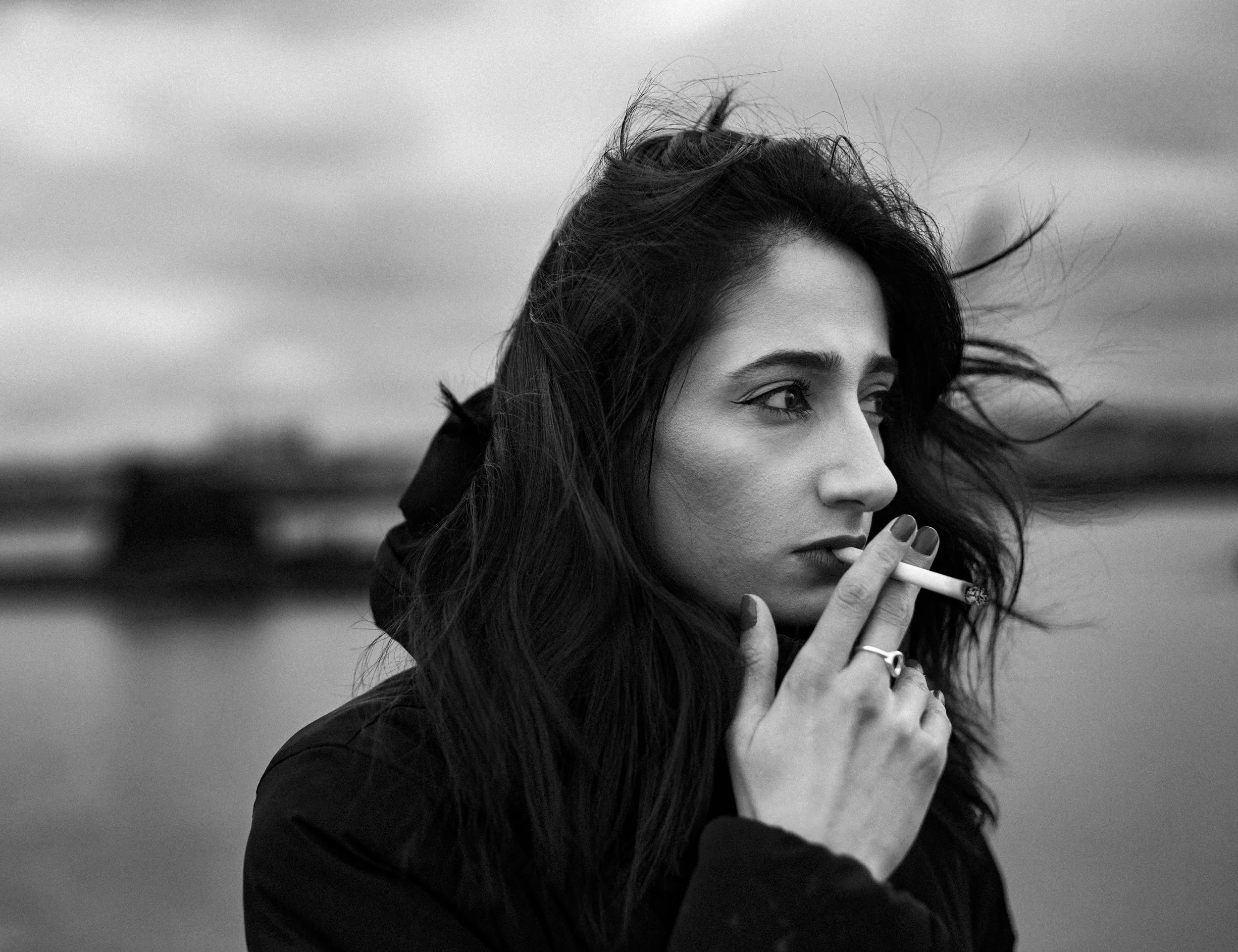 A black and white close-up image of a woman with wind in her hair, holding a cigarette, with an outdoor background.