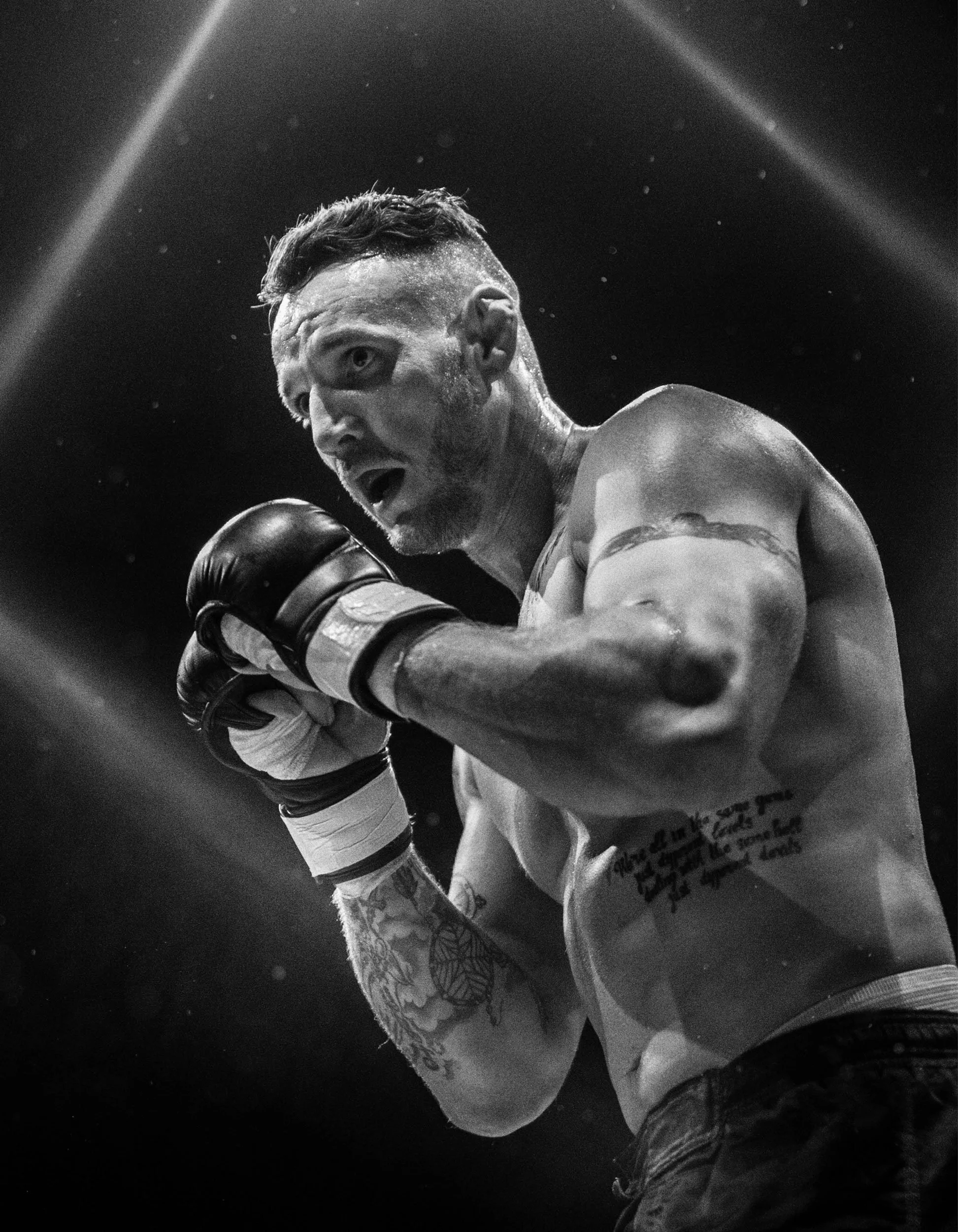 A shirtless male boxer with tattoos on his arm and chest, wearing boxing gloves, appears to be in a fighting stance inside a boxing ring, with a surprised or intense expression on his face.
