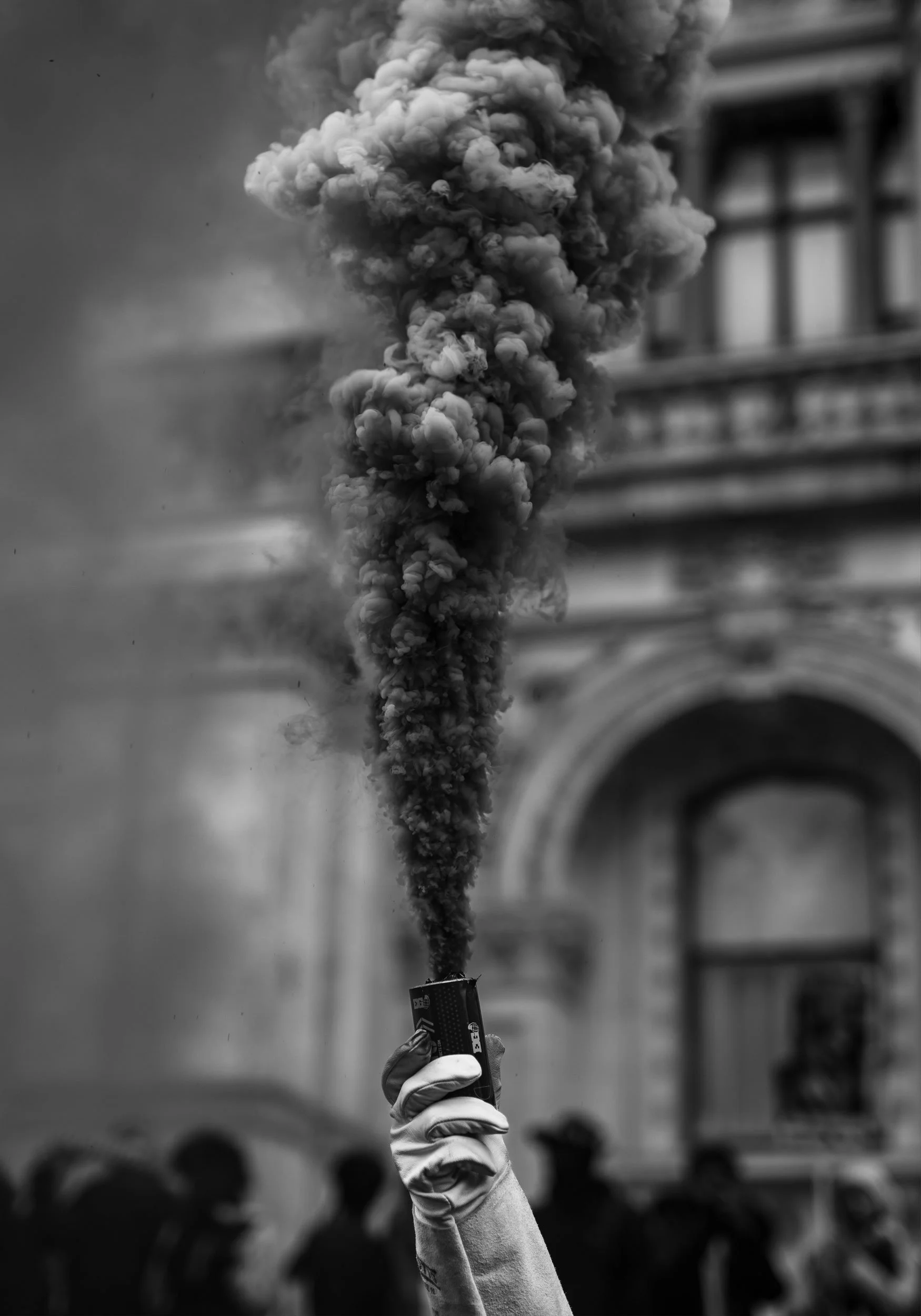 A black and white photo of a person holding a small smoke flare emitting a thick cloud of dark smoke in the air. In the background, blurred silhouettes of people are visible, with a building featuring arched windows and architectural details.