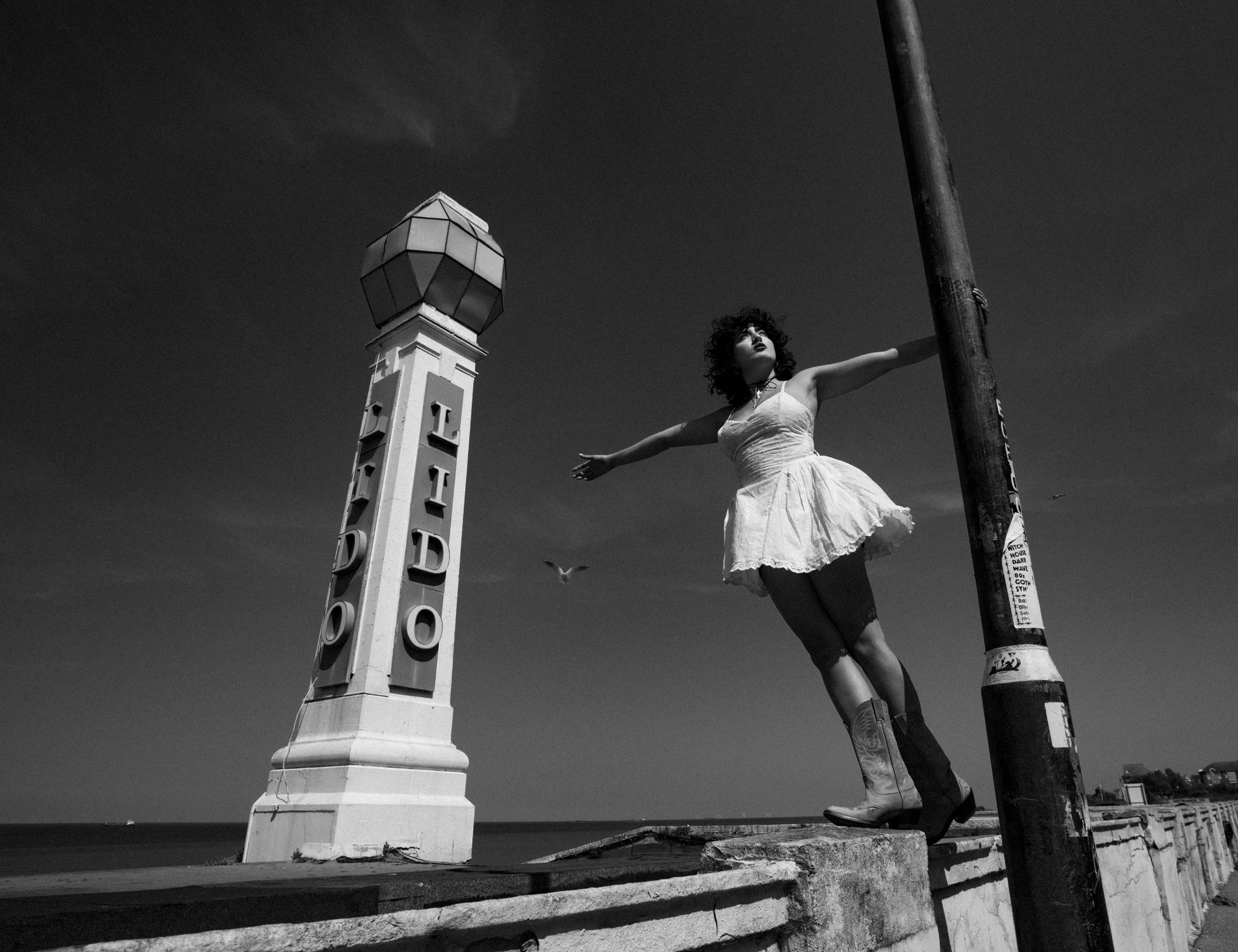 Yasmin Van Smith next to the Lido in Margate, Kent. Black and white photo of a woman in a dress with her arms outstretched, standing on a curb near a lamp post with a sign and a lighthouse-shaped structure next to her, and birds flying in the sky.