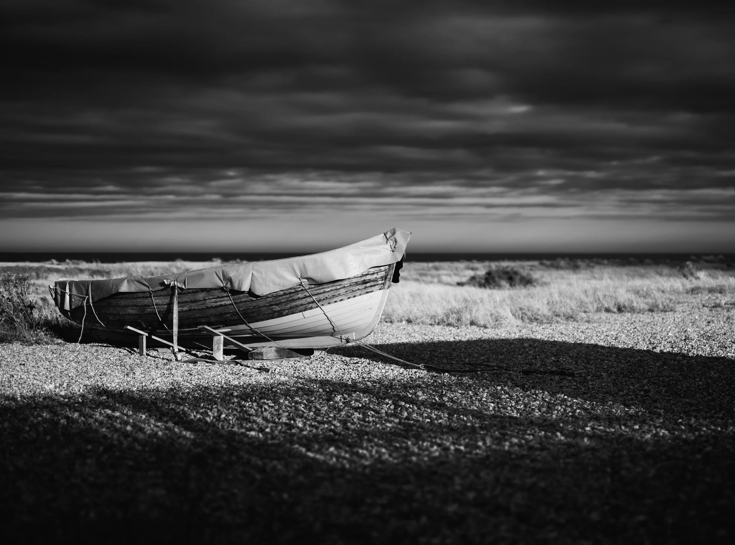 A small boat with a cover on it sits on a rocky beach under a cloudy sky in black and white.