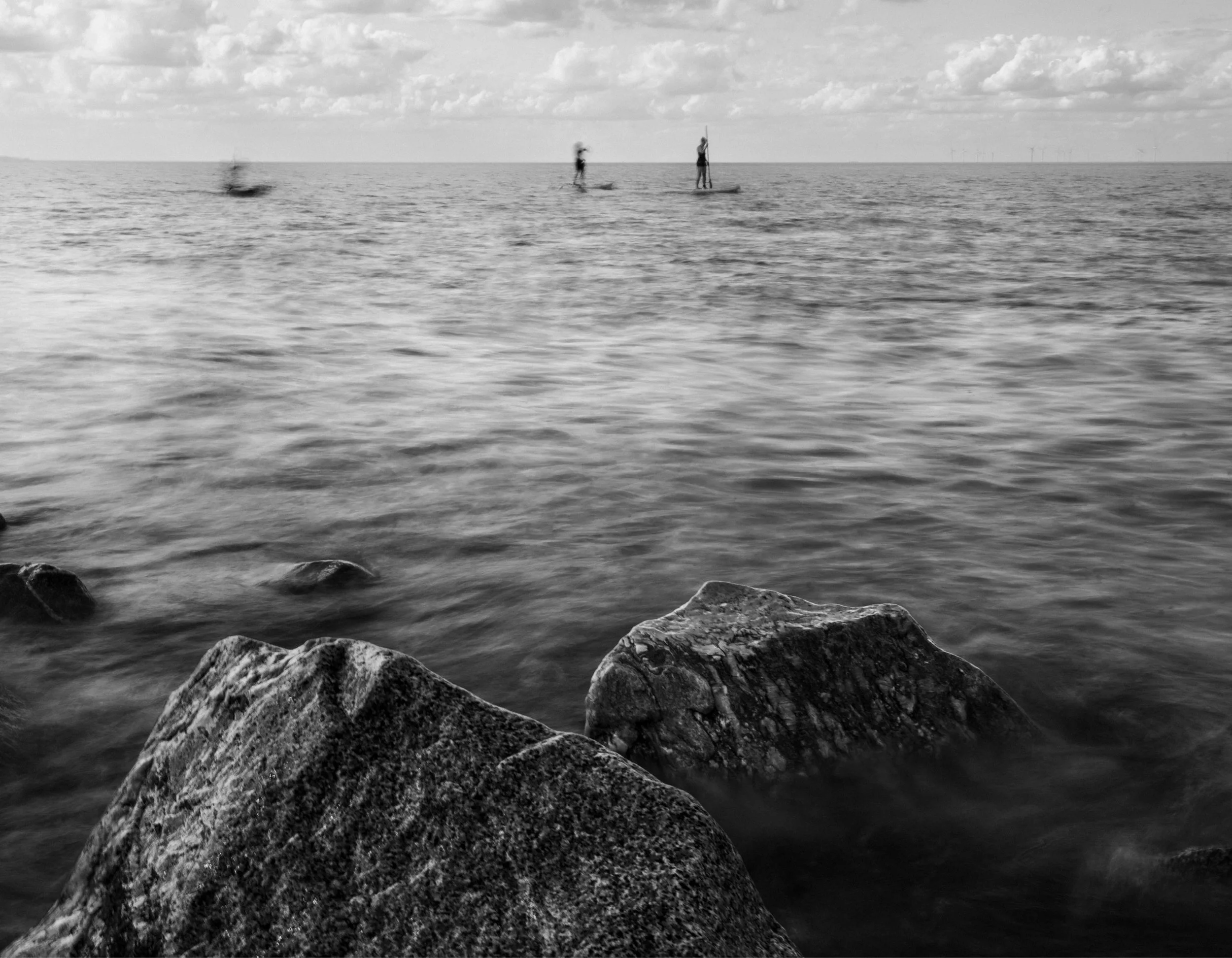 Black and white photo of a seascape with large rocks in the foreground, water in the middle, and three paddleboarders in the distance.