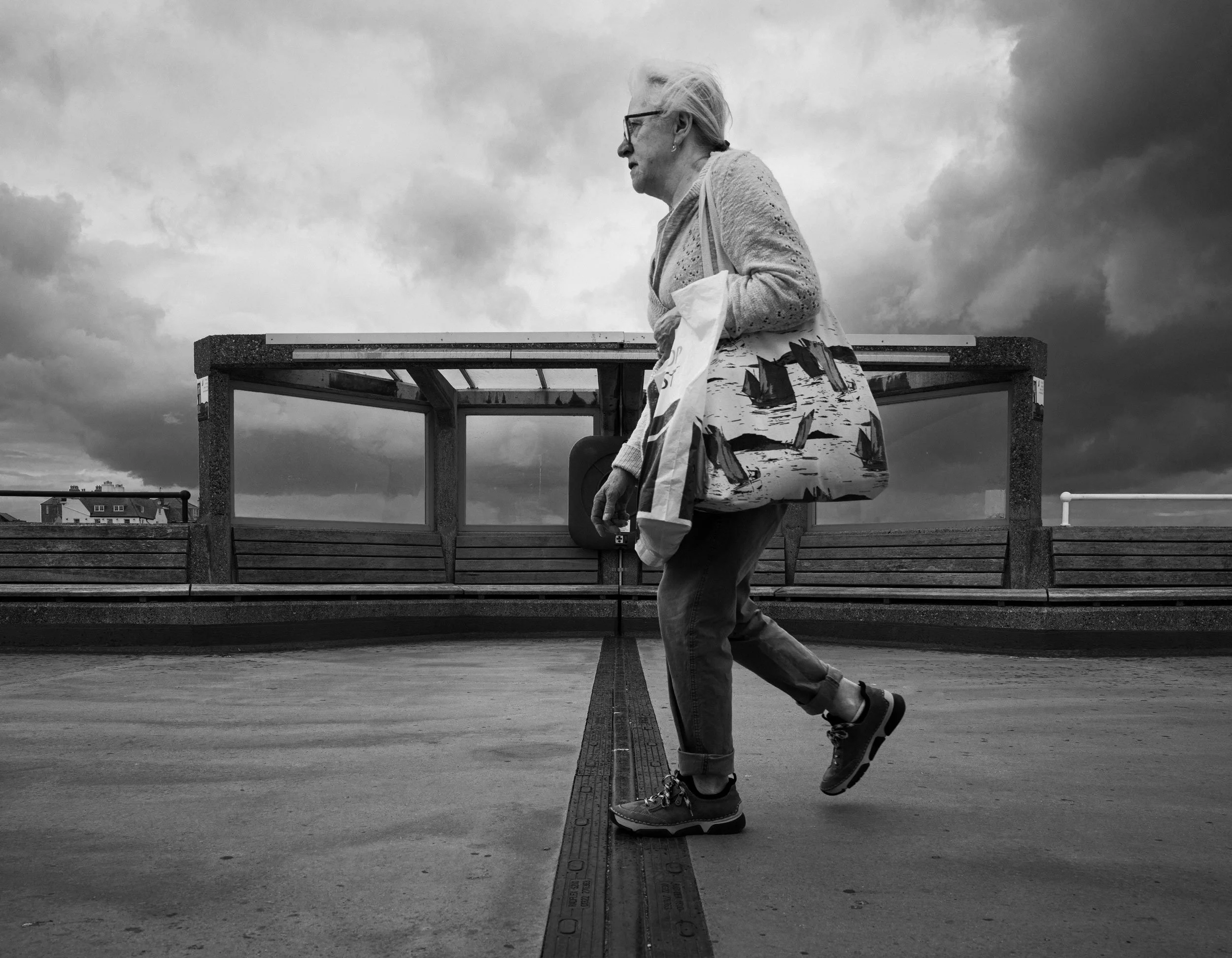 An elderly woman walking on a rooftop or parking structure with a cloudy sky in the background. She is carrying a tote bag with sailboat designs and holding a pair of sunglasses. The photo is in black and white.