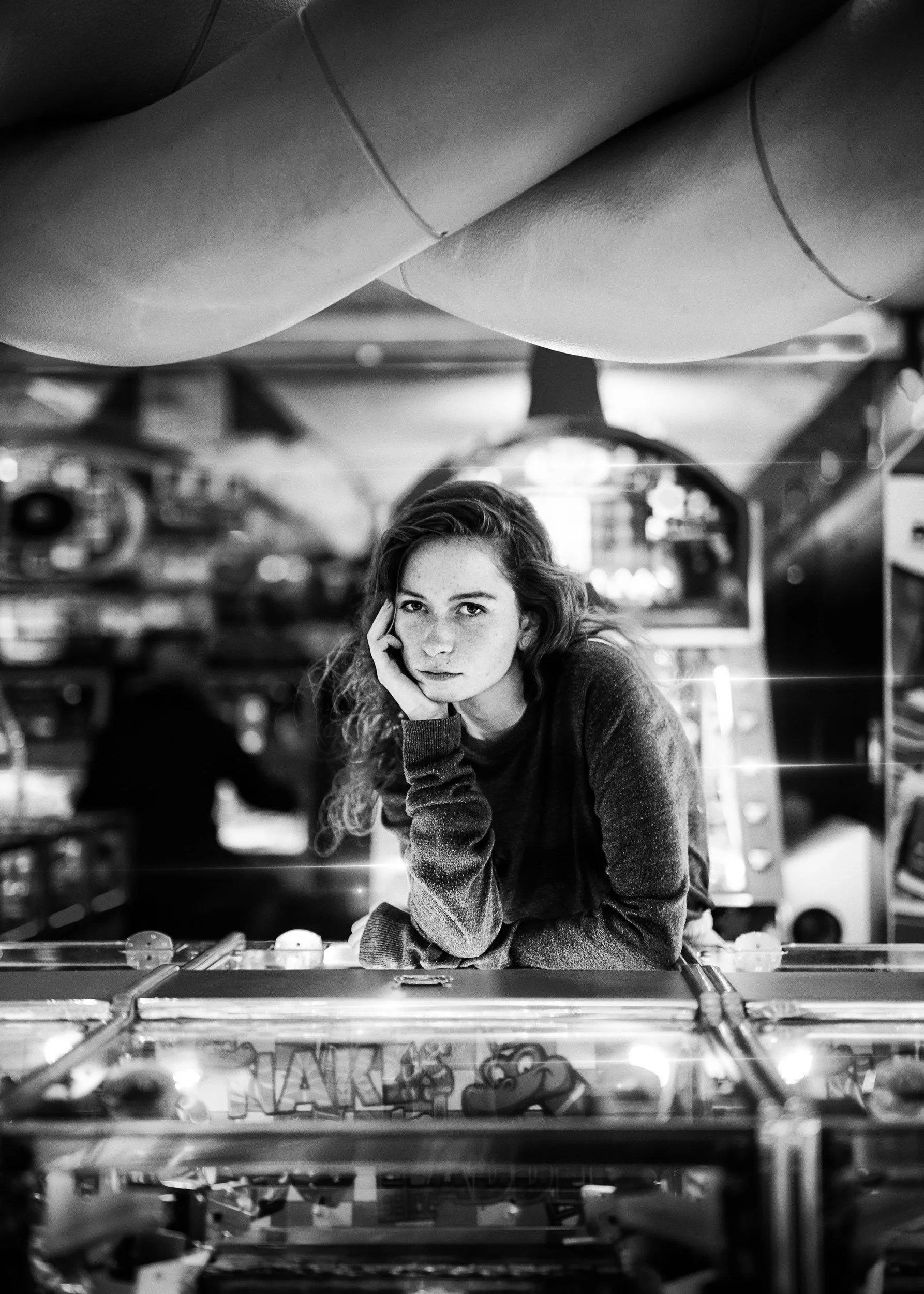 A young woman with curly hair and freckles leaning on her hand, sitting at a pinball machine in a game arcade, in black and white.