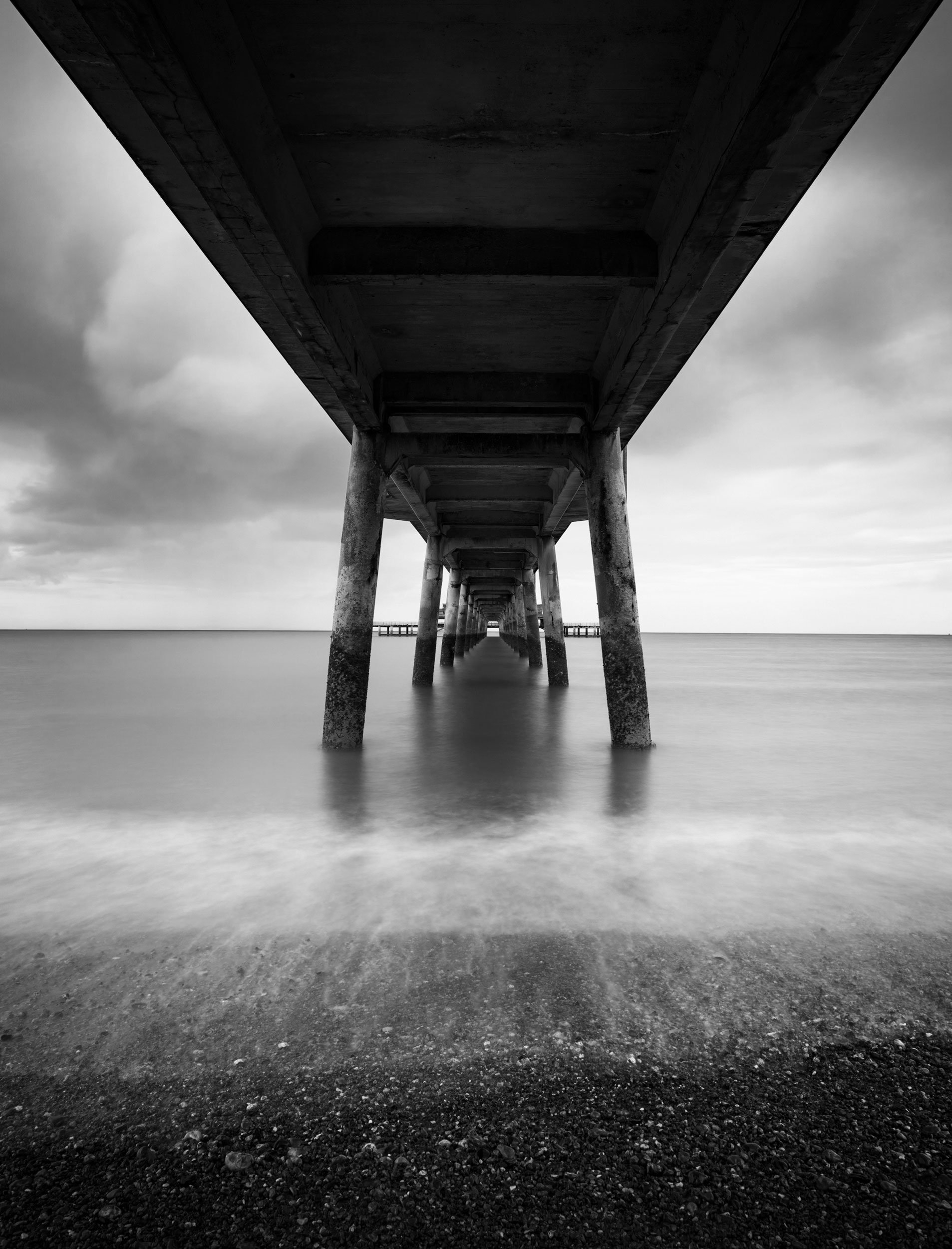 Underwater view of a pier with concrete pillars, captured in black and white, with calm water and cloudy sky in the background.