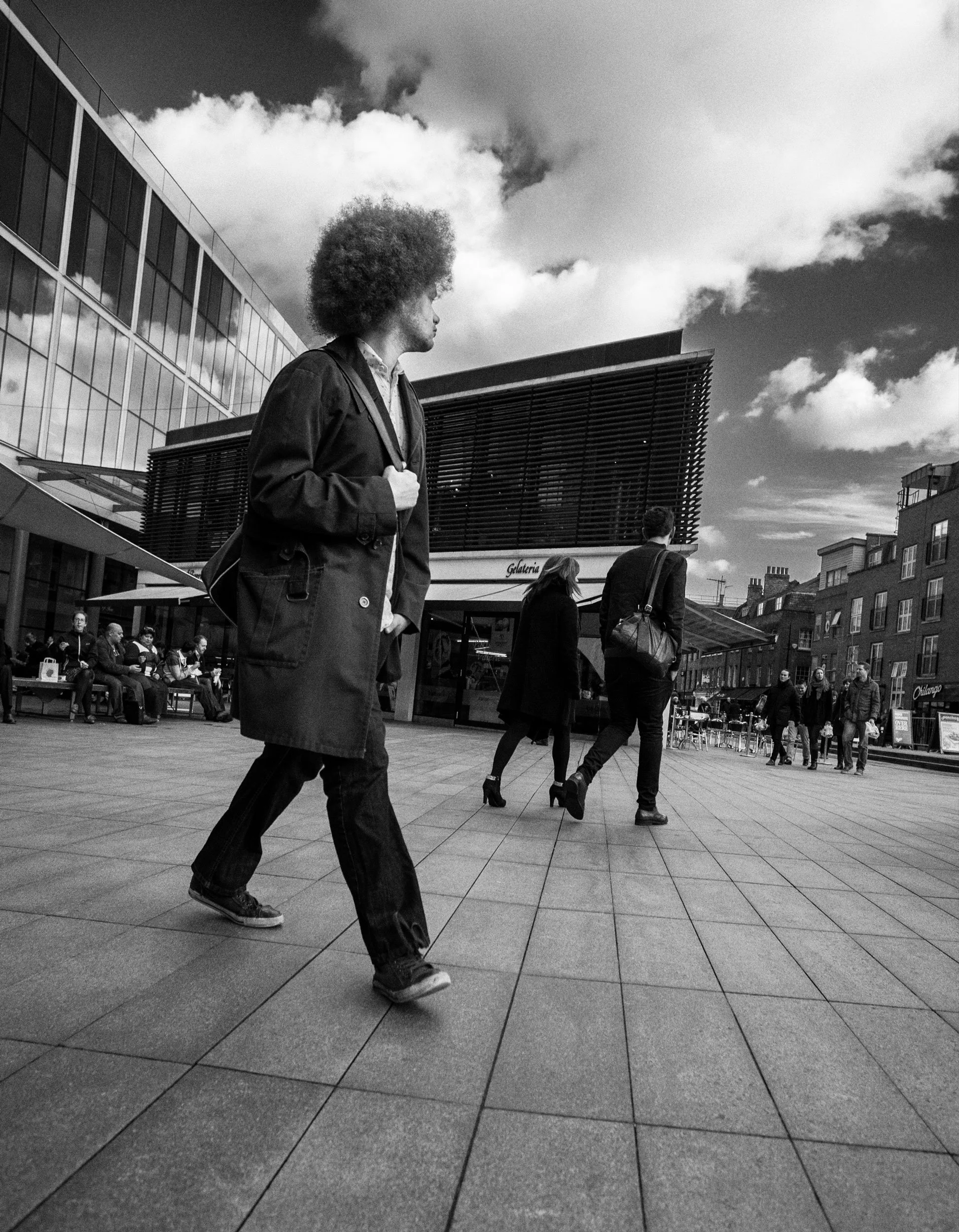 Black and white photo of people walking outdoors in an urban area near a modern glass building and a structure with horizontal slats, with a cloudy sky overhead.