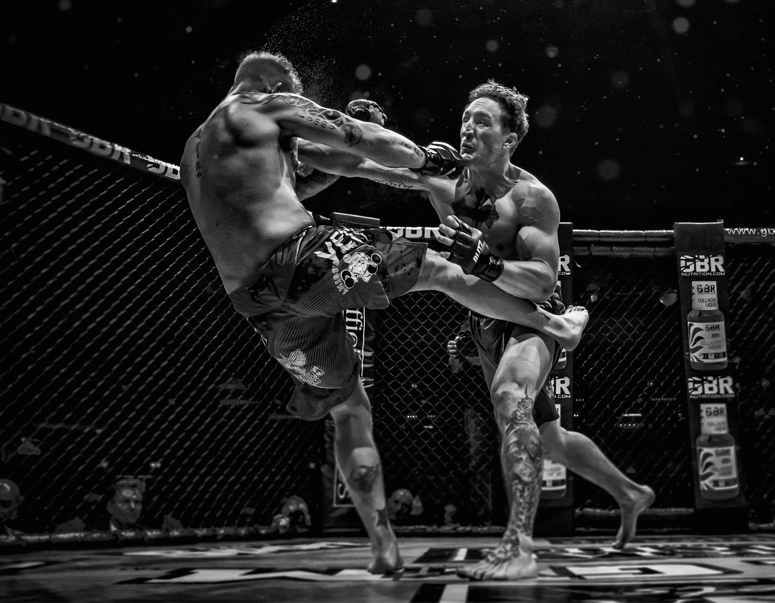 Two mixed martial arts fighters engaged in a fight inside an octagon. One fighter is delivering a punch while the other is blocking. The scene is intense with sweat and includes a visible audience and sponsor logos.