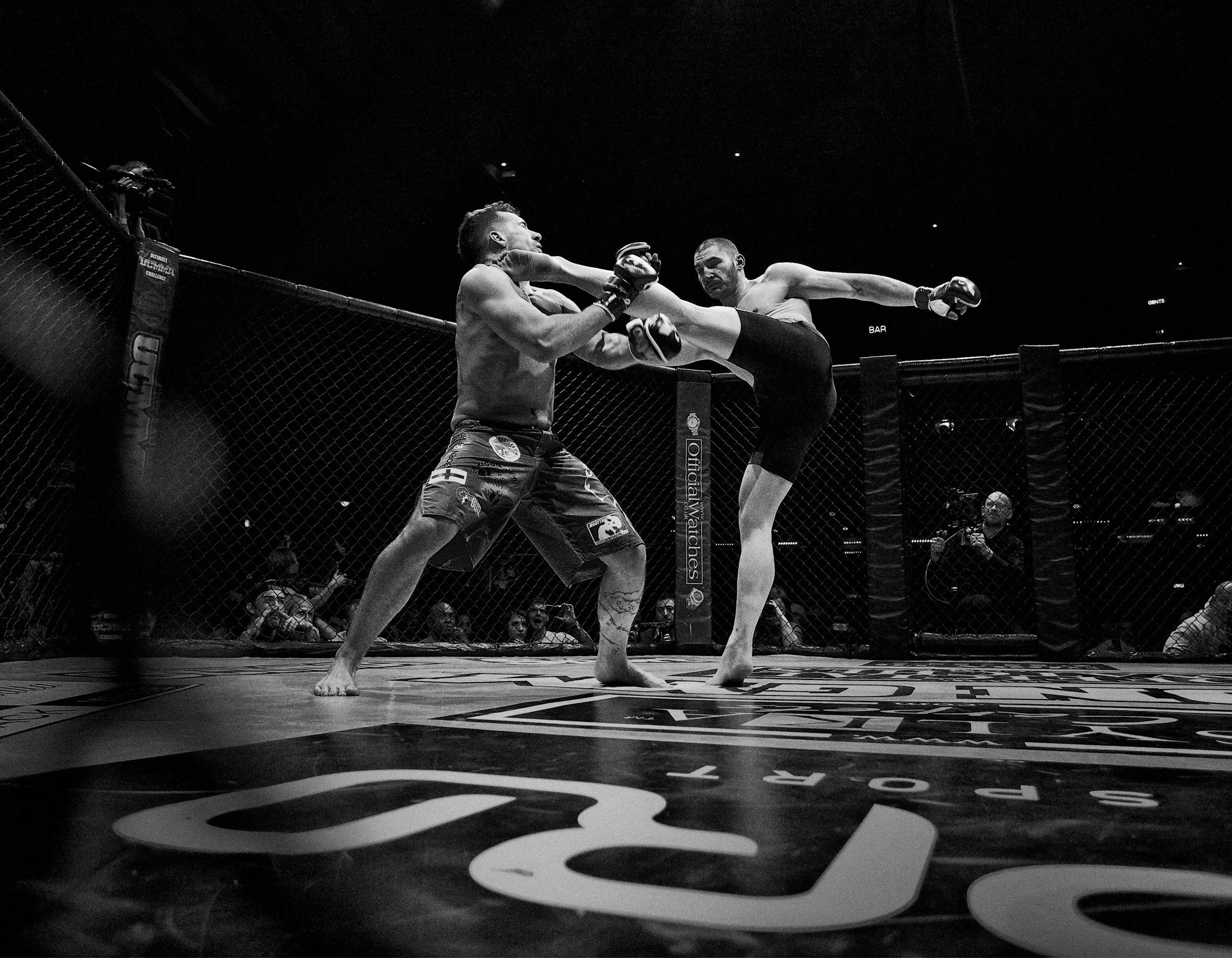 Two mixed martial arts fighters inside a cage, one delivering a high kick to the other's face, with spectators watching in the background.