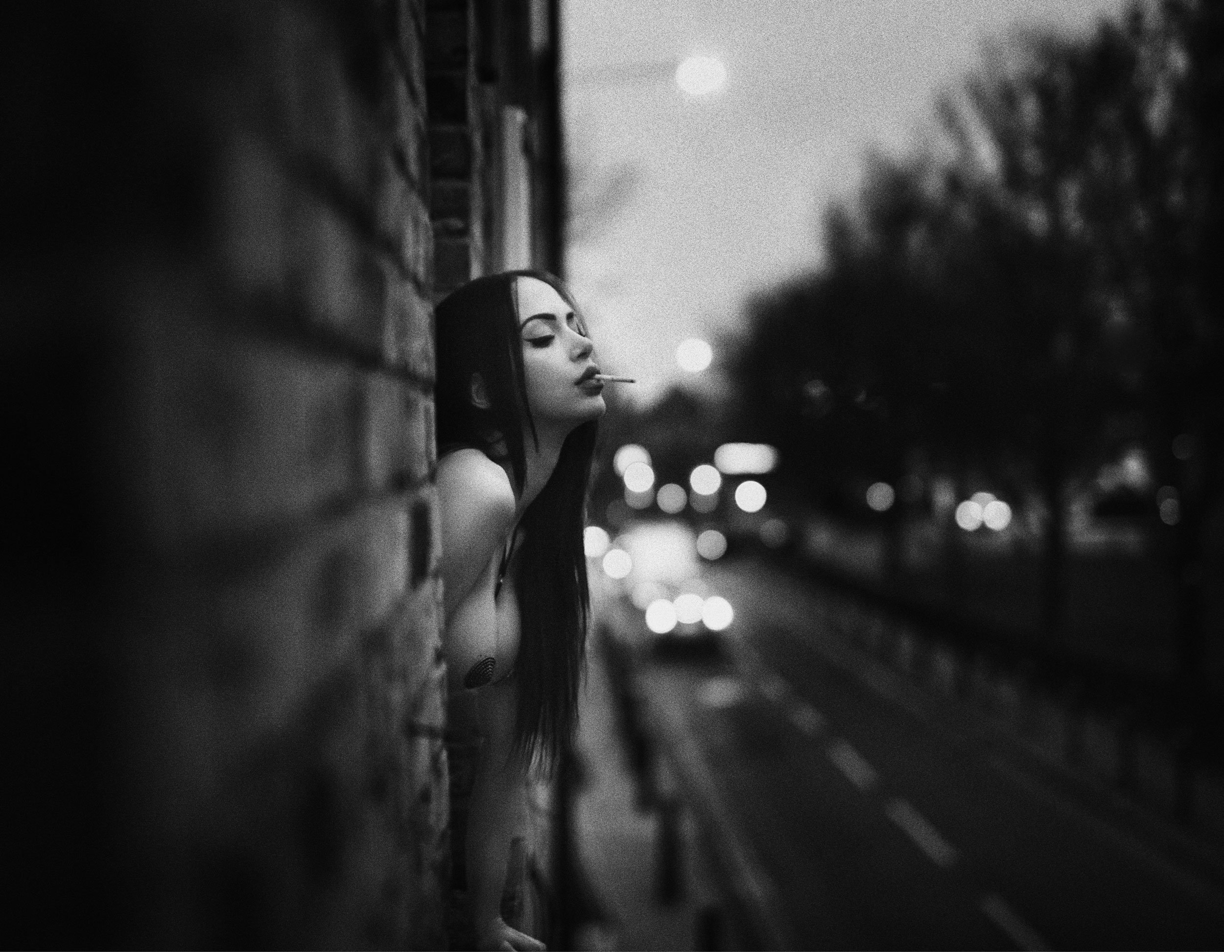 A woman with dark hair and makeup smoking a cigarette outdoors at night, leaning against a brick wall with blurred city lights in the background.