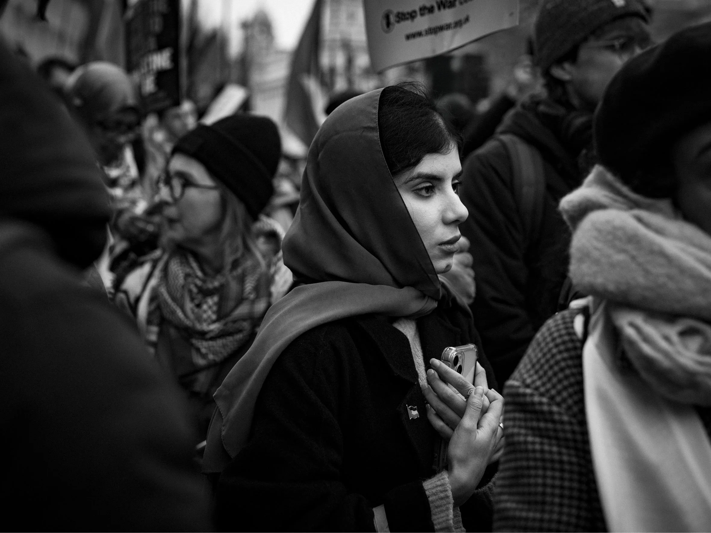 A black and white photo of a young woman with dark hair and a headscarf holding a small object close to her chest during a protest or gathering, surrounded by other diverse individuals.