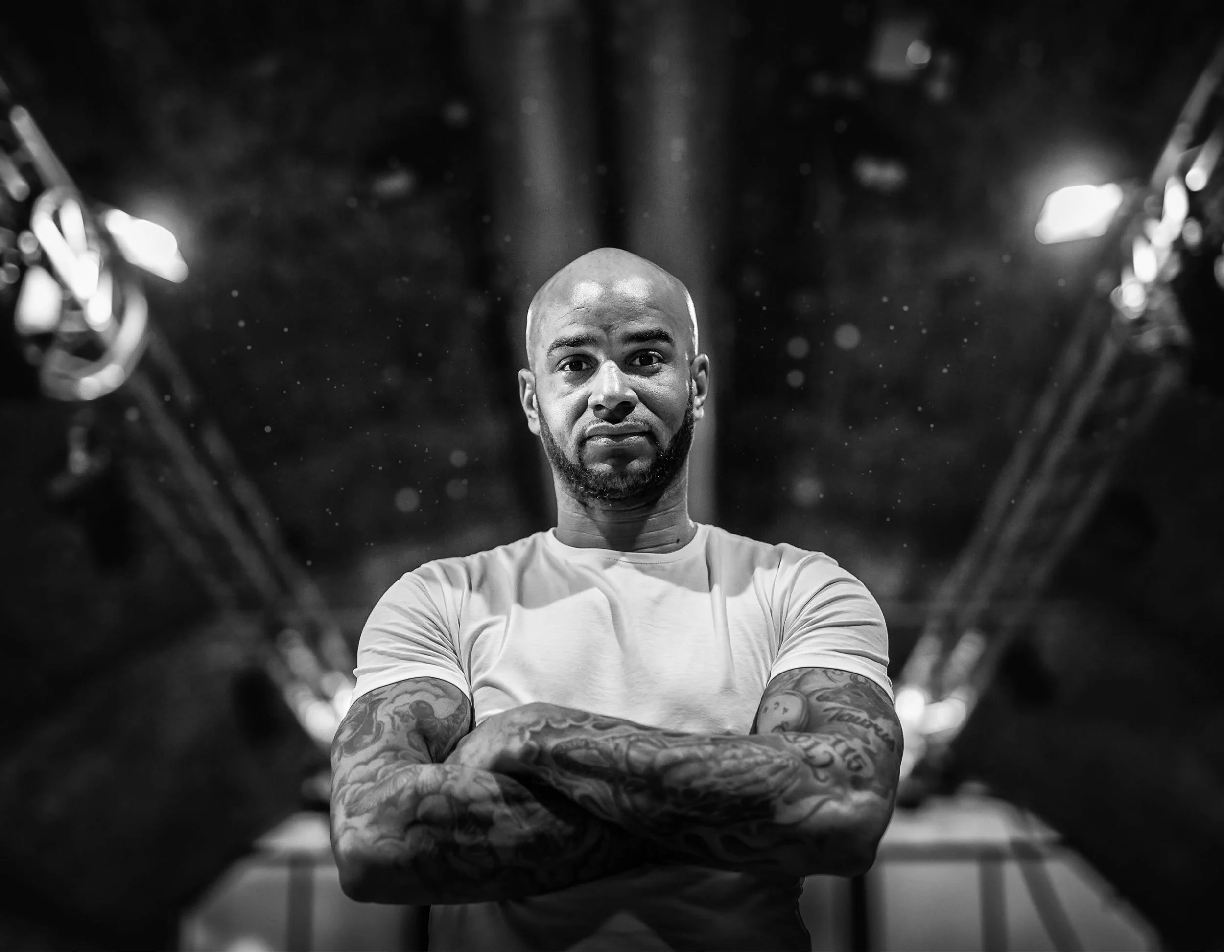 Black and white photo of a bald man with tattoos on his arms, crossing his arms and looking at the camera in a gym or studio with lights and equipment in the background.