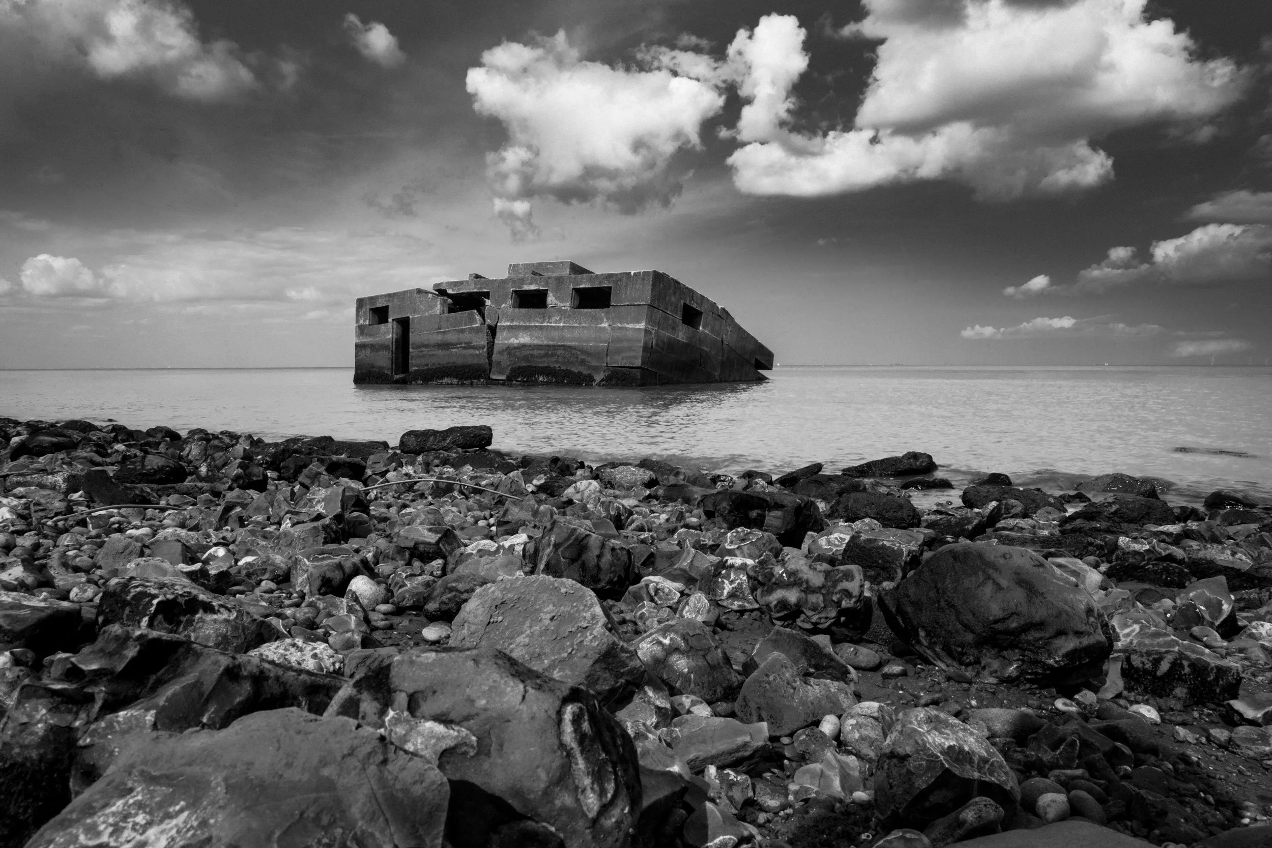 Black and white photo of a shipwreck partially submerged in water near a rocky shoreline with a cloudy sky.