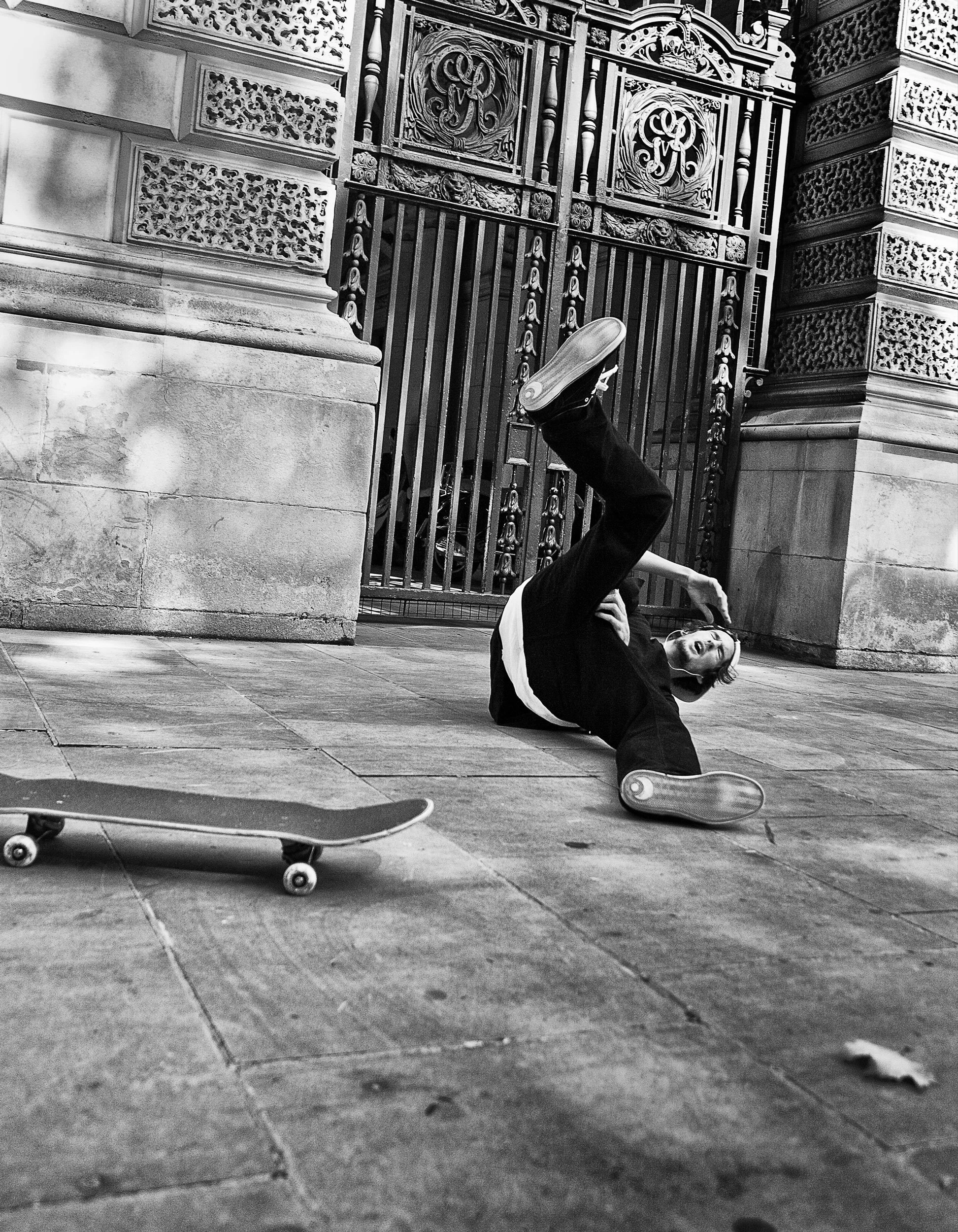 A person falling down on the sidewalk with a skateboard nearby, in front of a decorative iron gate.