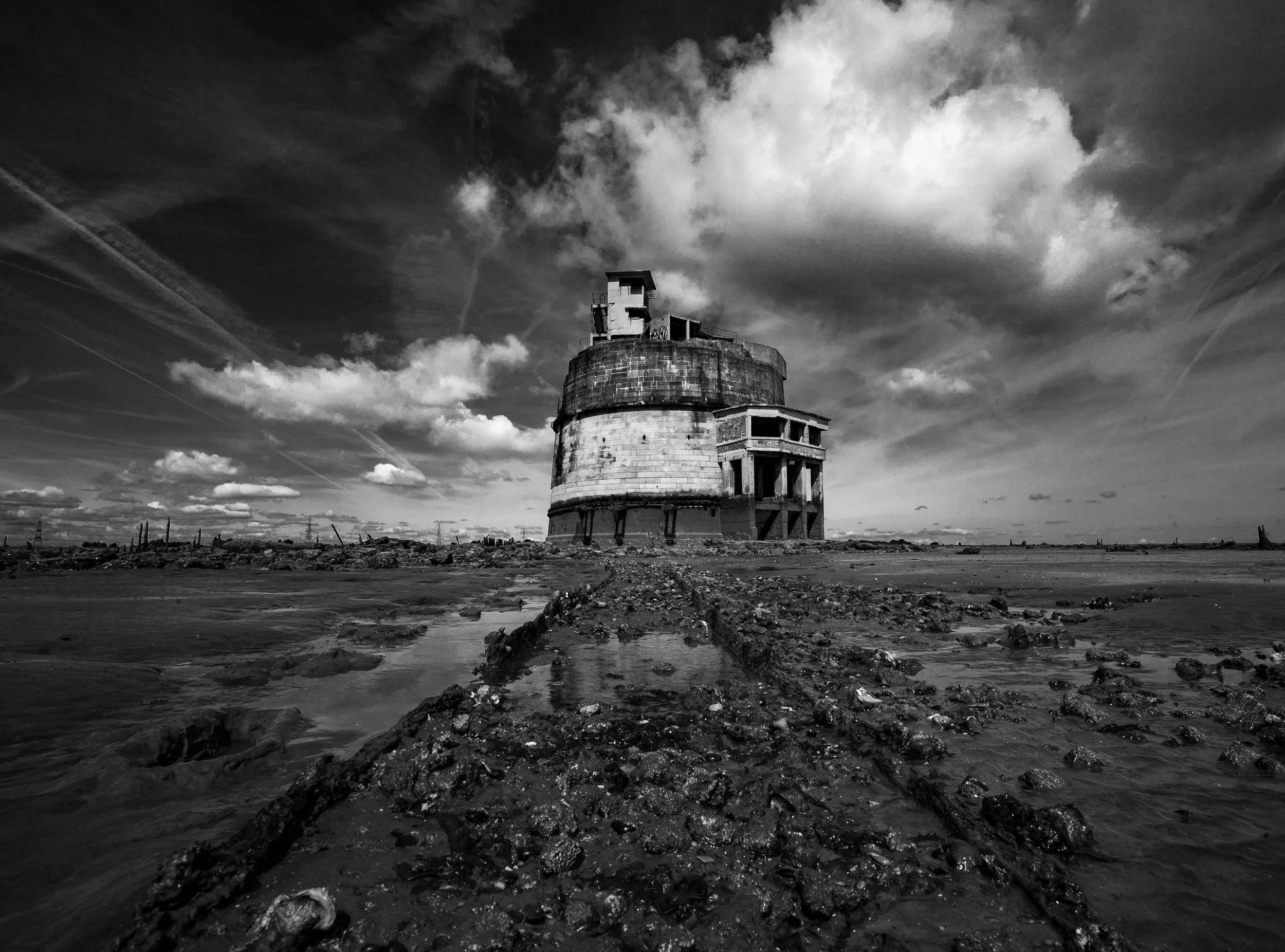 Black and white photo of an abandoned, large, circular concrete structure on a muddy, rocky landscape under a cloudy sky.