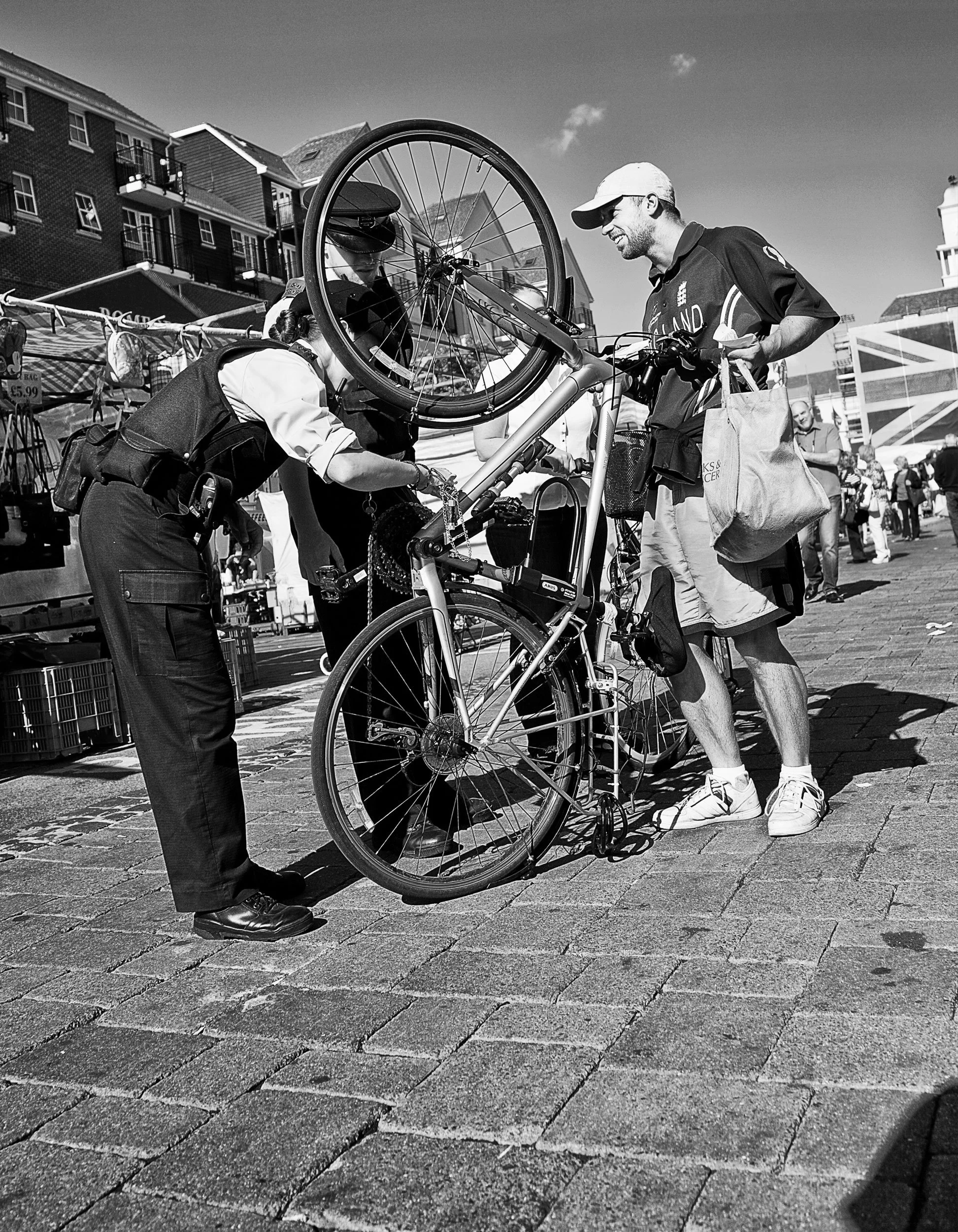 A man with a bicycle being checked by police officers at an outdoor market or festival, with stalls and people in the background.
