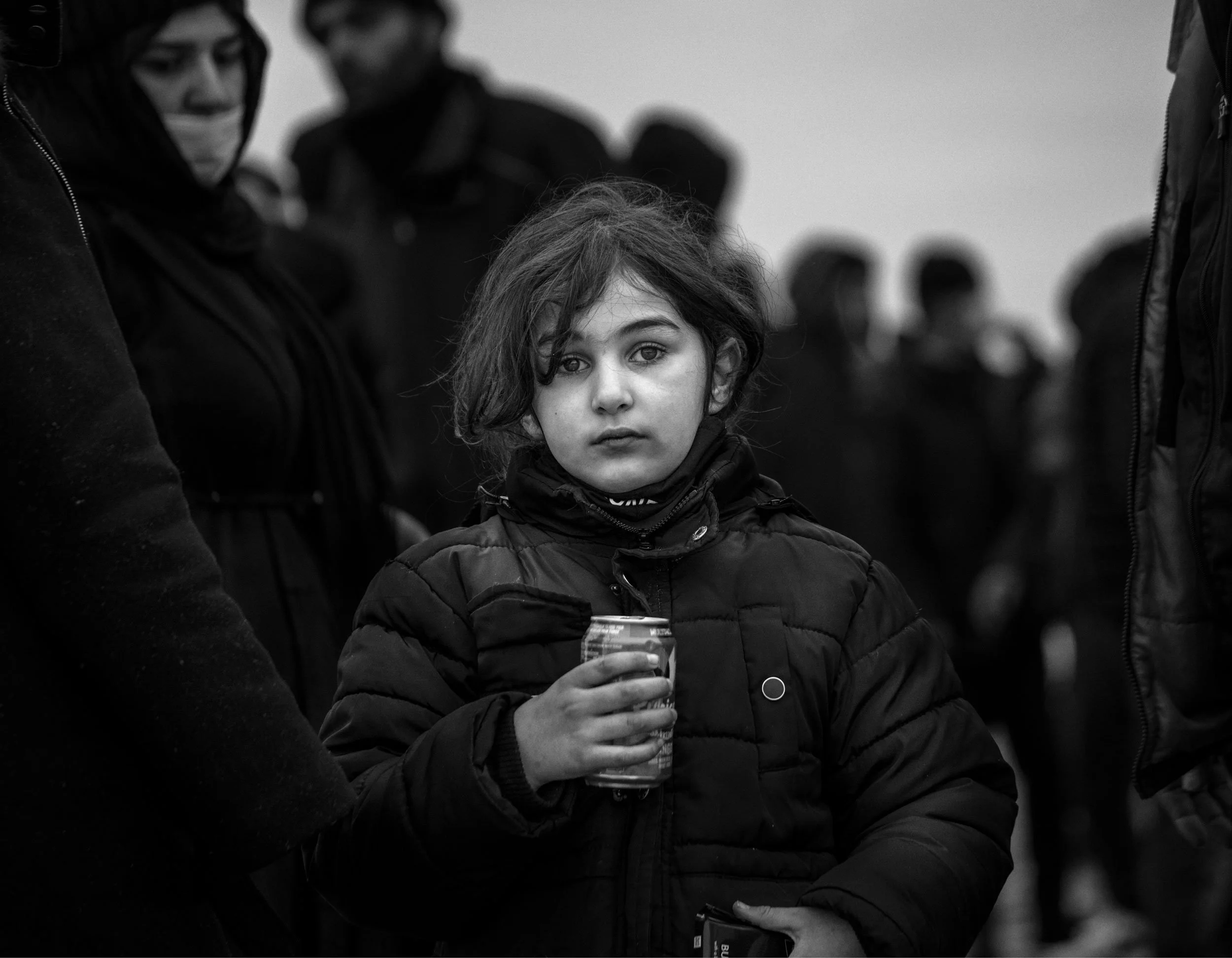 A young girl with dark hair and expressive eyes, wearing a puffy jacket, holding a drink in her right hand, standing among a crowd of people outdoors in a grayscale image.