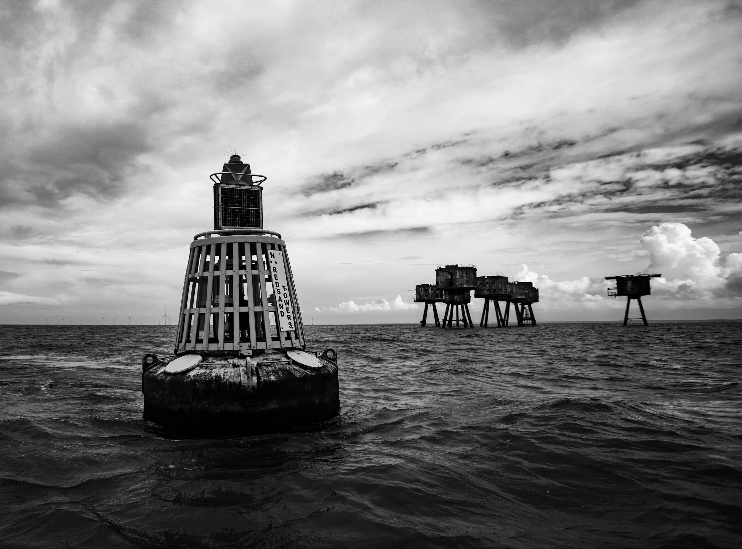 Black and white photo of offshore oil rigs in the ocean, with a buoy marked "N. RED SAND TOWERS" in the foreground. Overcast sky with scattered clouds. Maunsell Forts