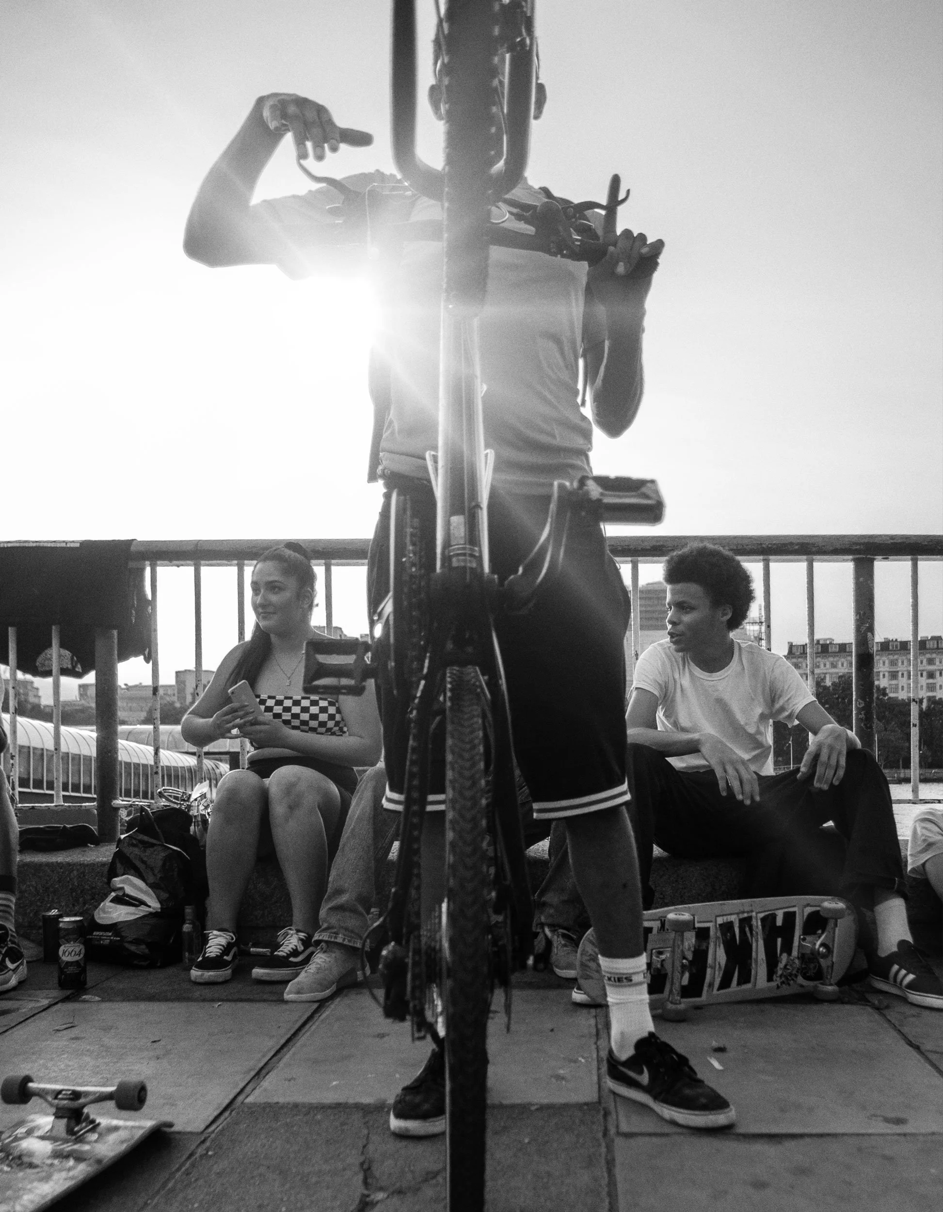 A black and white photo of a young man standing with a bicycle on a bridge, with two young women sitting on the ground behind him. One woman is looking at her phone, and the other is looking to the side. There are skateboards and backpacks nearby.
