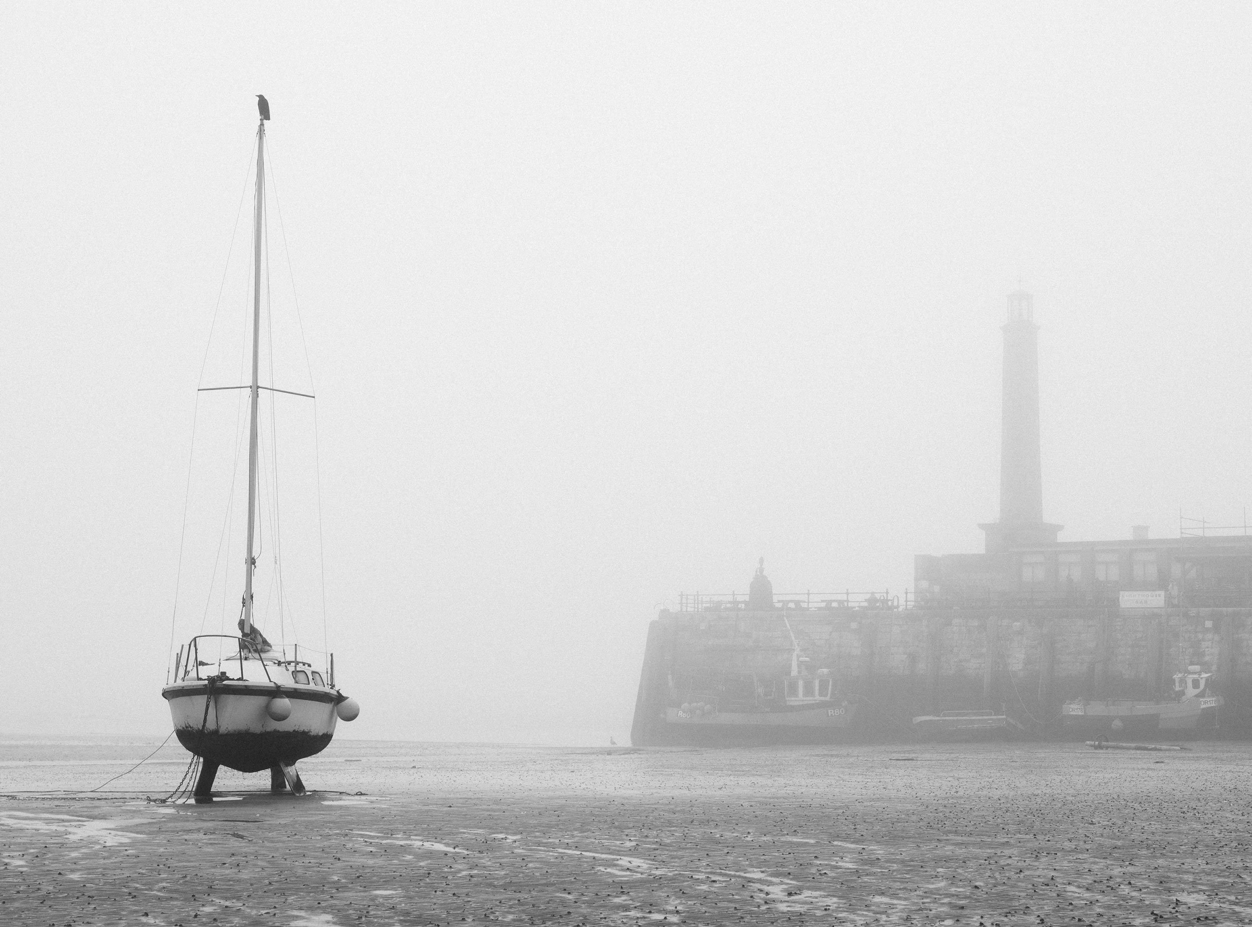 A sailboat resting on the sandy shore in front of a foggy harbor scene, with a lighthouse and ships faintly visible in the background. Margate beach on a foggy day.