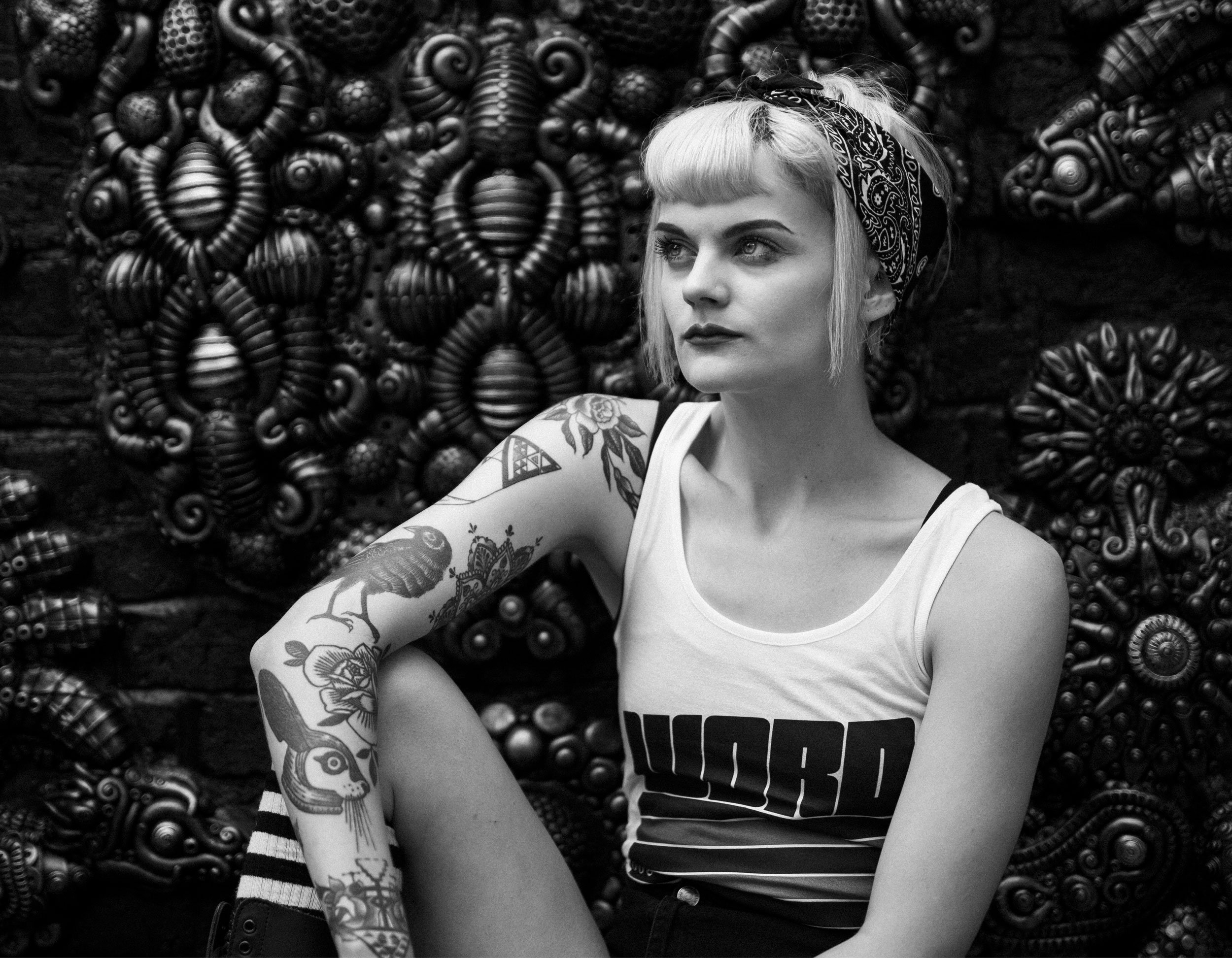 A young woman with short blonde hair, tattoos on her arm, wearing a sleeveless shirt and a bandana on her head, sitting against an artistic backdrop of various textured, decorated objects.