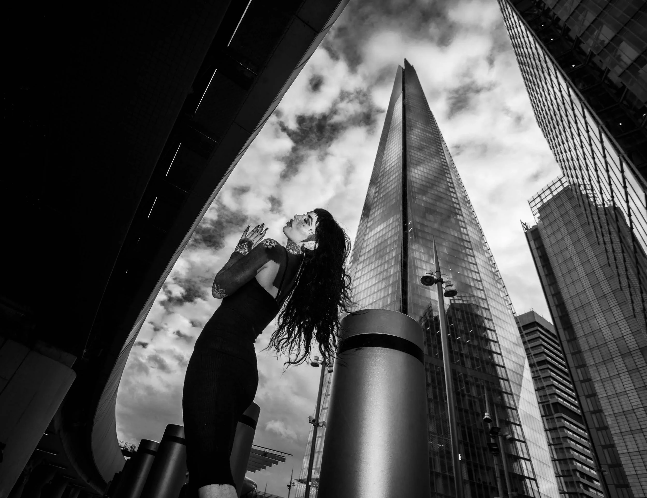 A woman with tattoos and tattoos on her face, dressed in black, stands in an urban setting with modern glass buildings around her. The photo is black and white, taken from a low angle, capturing the woman and tall skyscrapers beneath a cloudy sky.